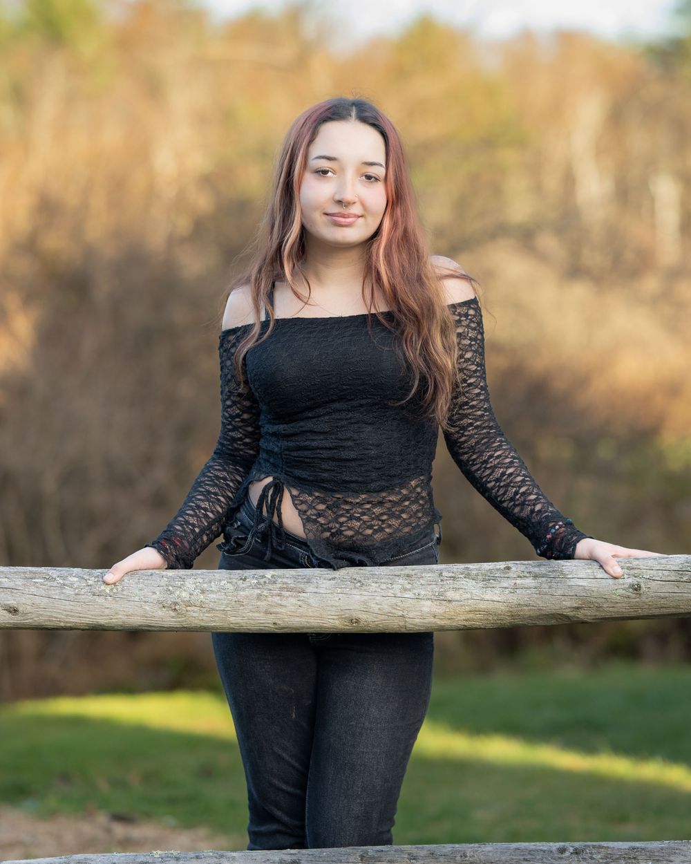 Young woman in black off-shoulder lace top and dark jeans posing outdoors by a wooden fence rail.