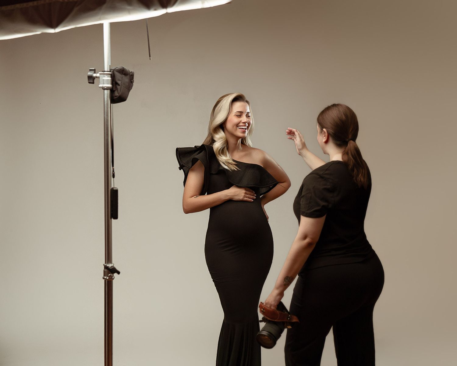 Pregnant woman in black maternity outfit poses with stylist during professional studio photoshoot on beige background.