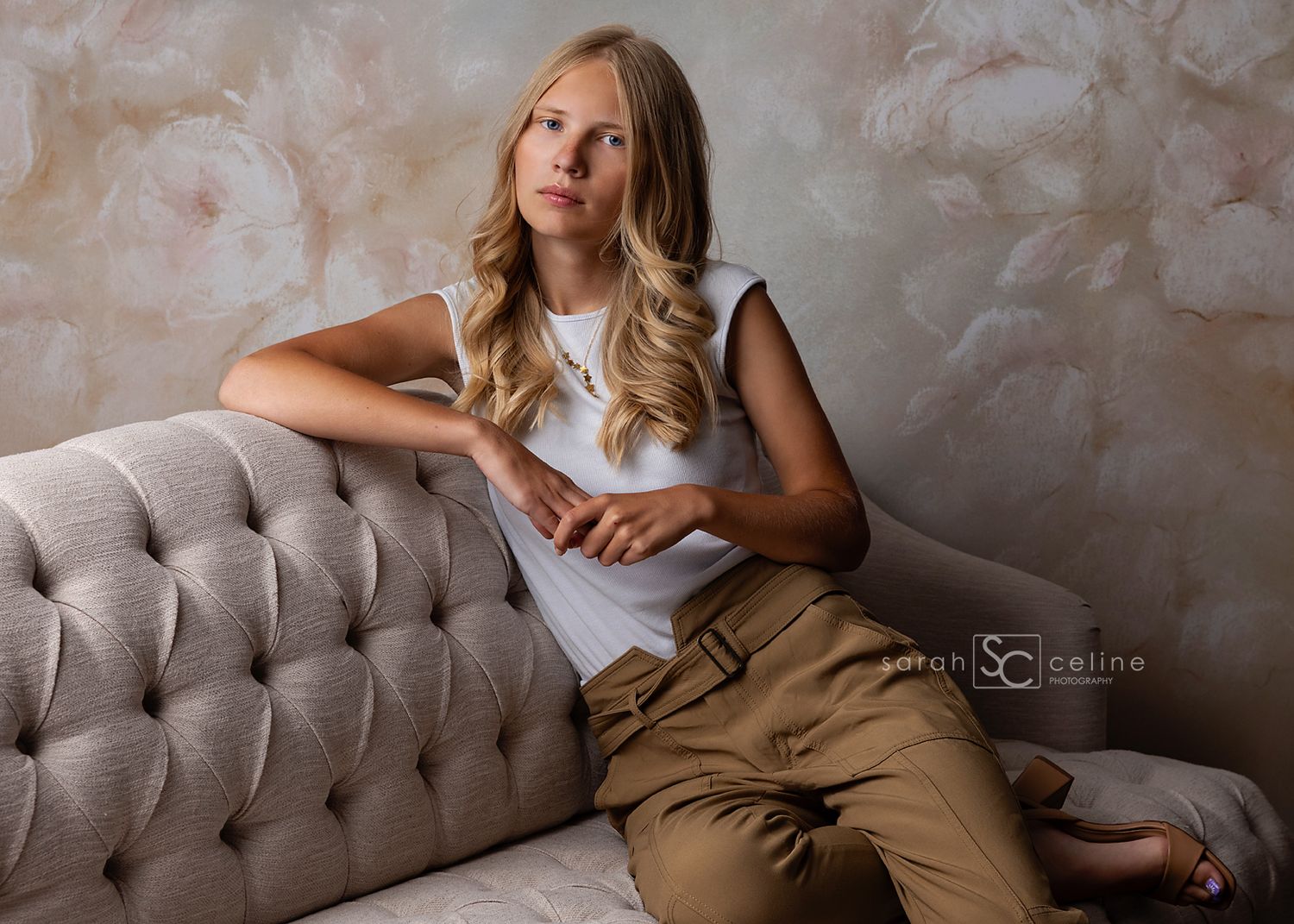 Teen girl in white top and tan pants poses on tufted sofa against floral backdrop in professional portrait.