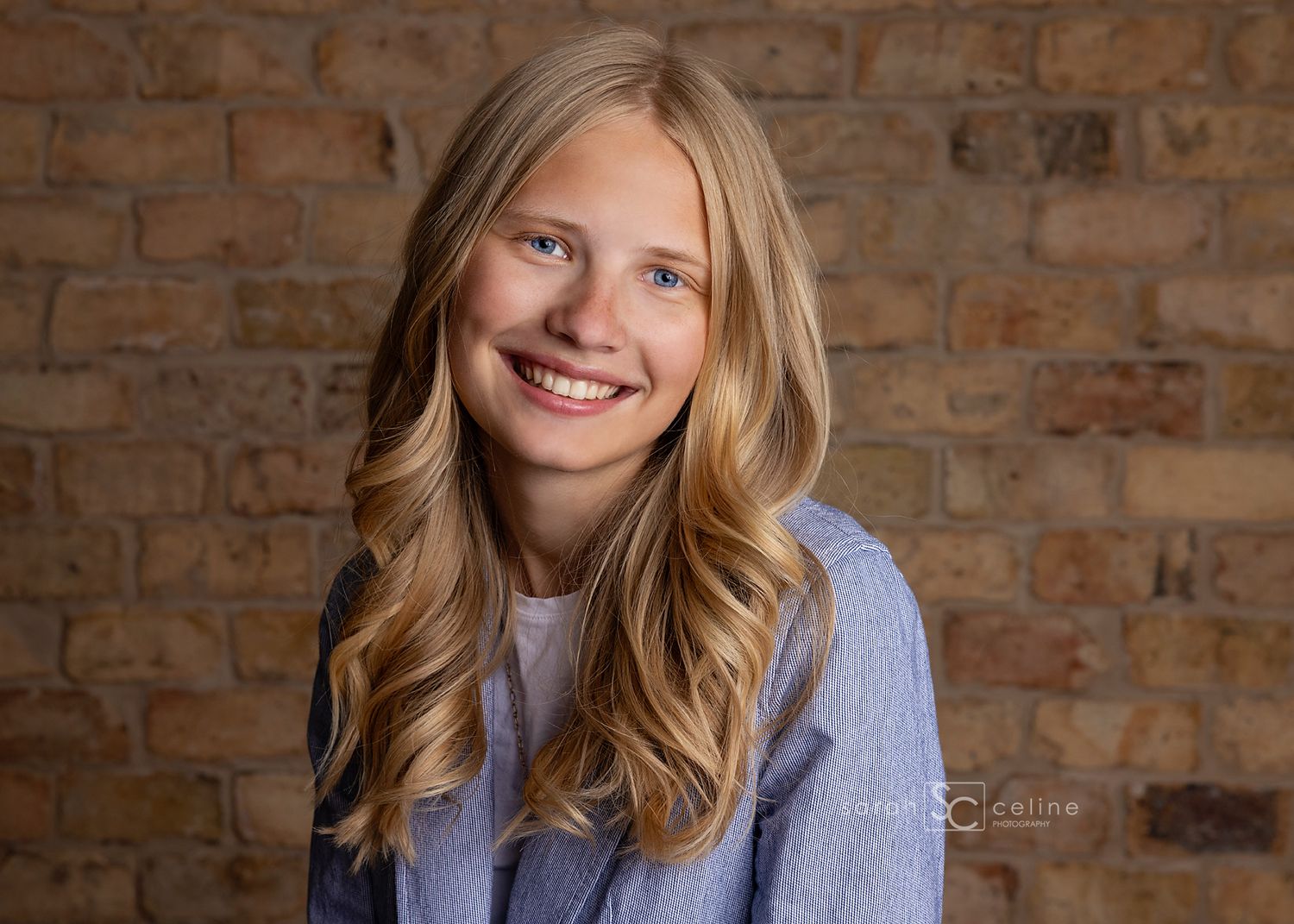 Smiling teenage girl with long blonde hair poses in front of a brick wall in a blue striped shirt.