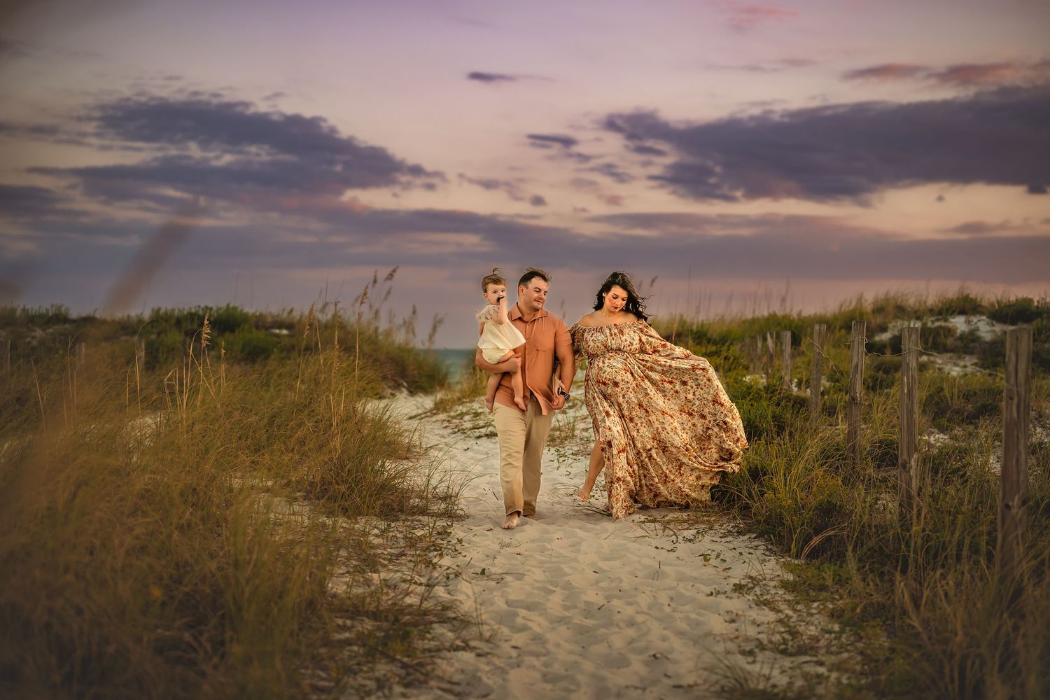 Family of three walking on sandy beach path at sunset, woman in floral gown, man holding toddler, golden dunes backdrop.