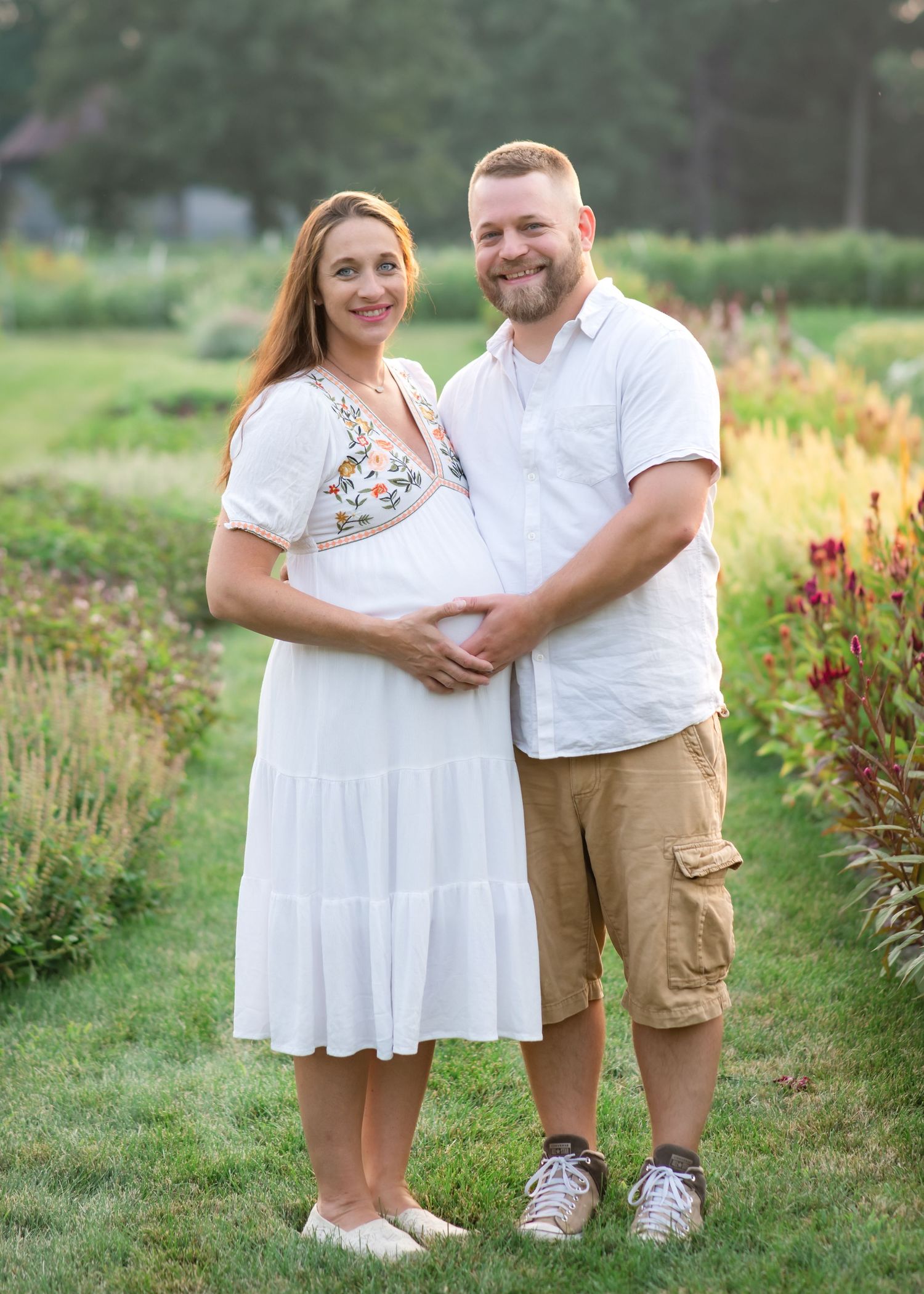 Smiling couple poses in a garden, woman in white dress with baby bump, man in white shirt and khaki shorts.
