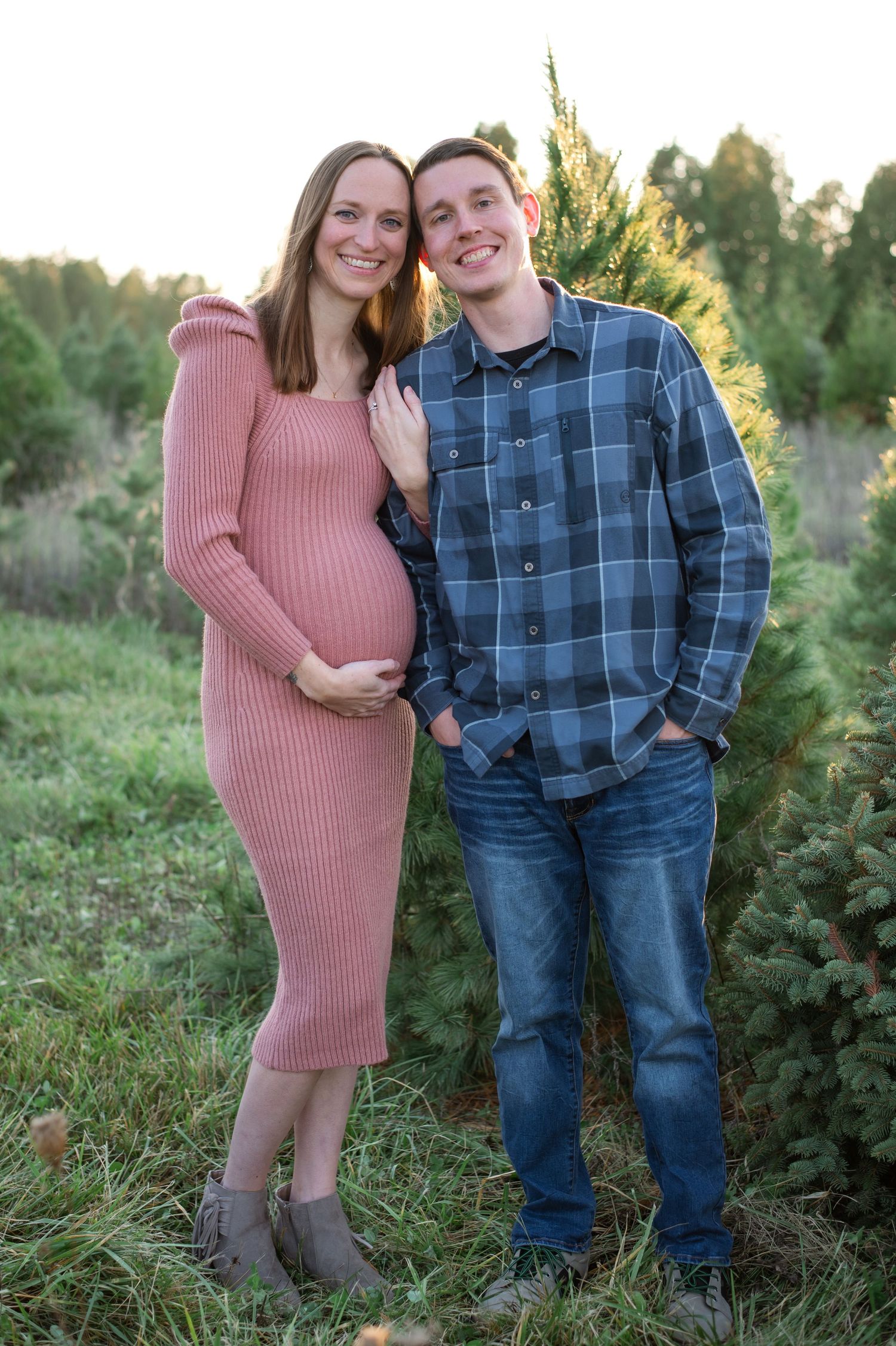 Smiling pregnant couple posing outdoors at a Christmas tree farm, woman in pink dress, man in plaid shirt.