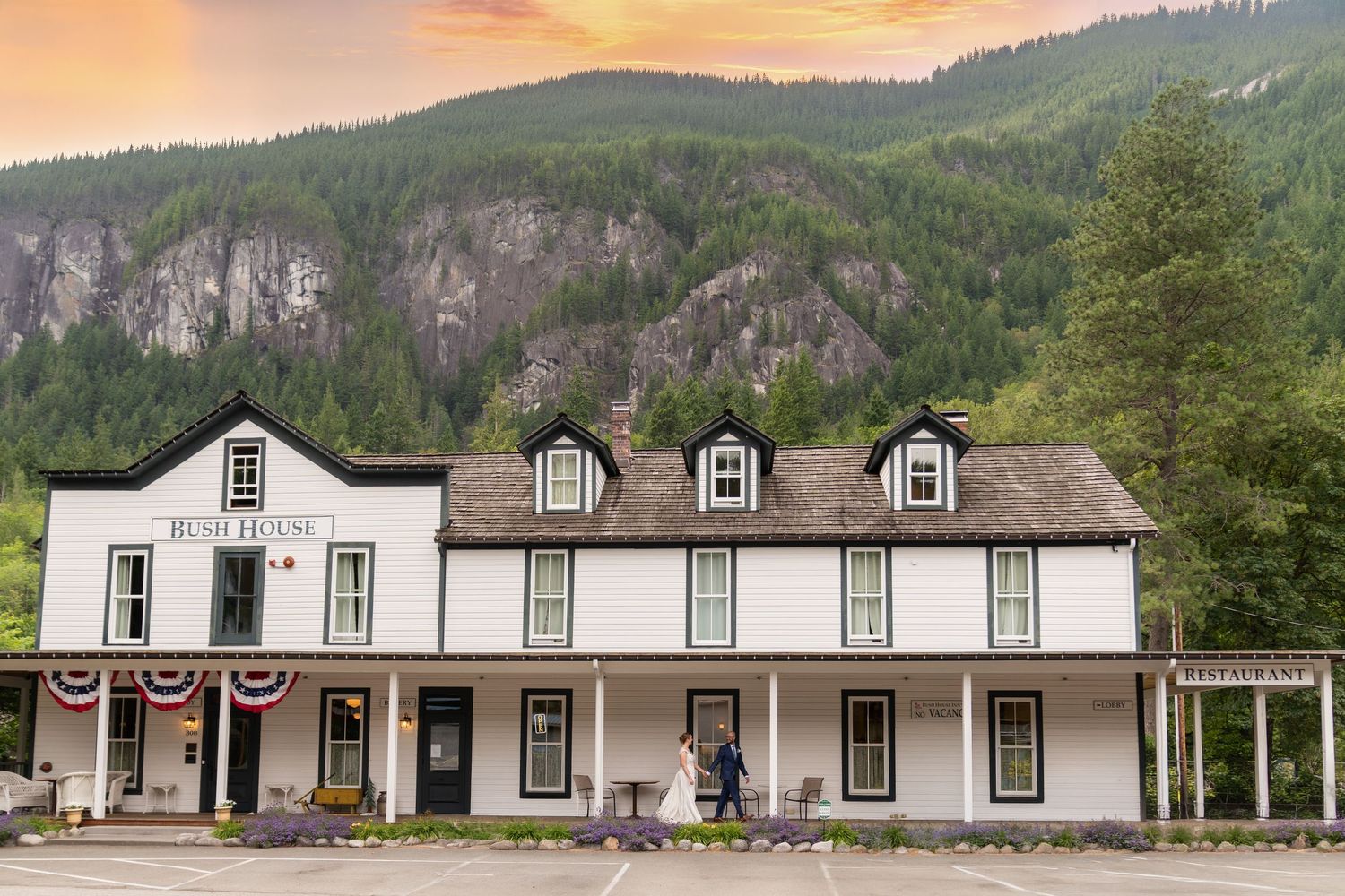 Historic Bush House hotel with white facade, patriotic bunting, mountain backdrop, and wedding couple at entrance.
