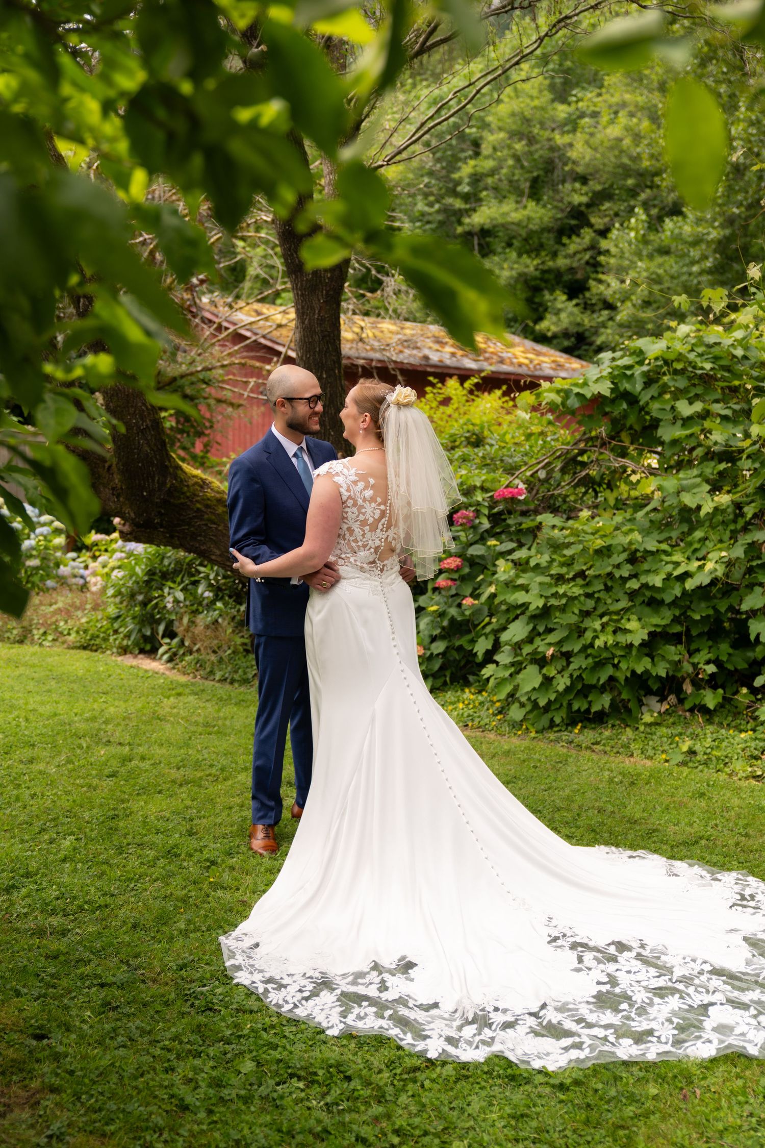 Bride in lace wedding gown with veil and groom in navy suit share a romantic moment in a lush garden setting.