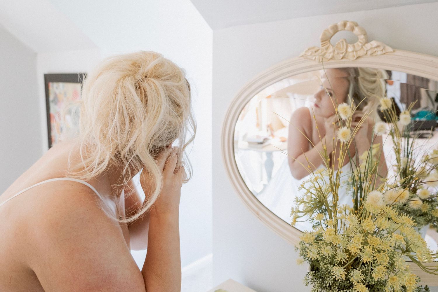 Blonde woman applying makeup while looking into an ornate white mirror with flowers in the foreground.