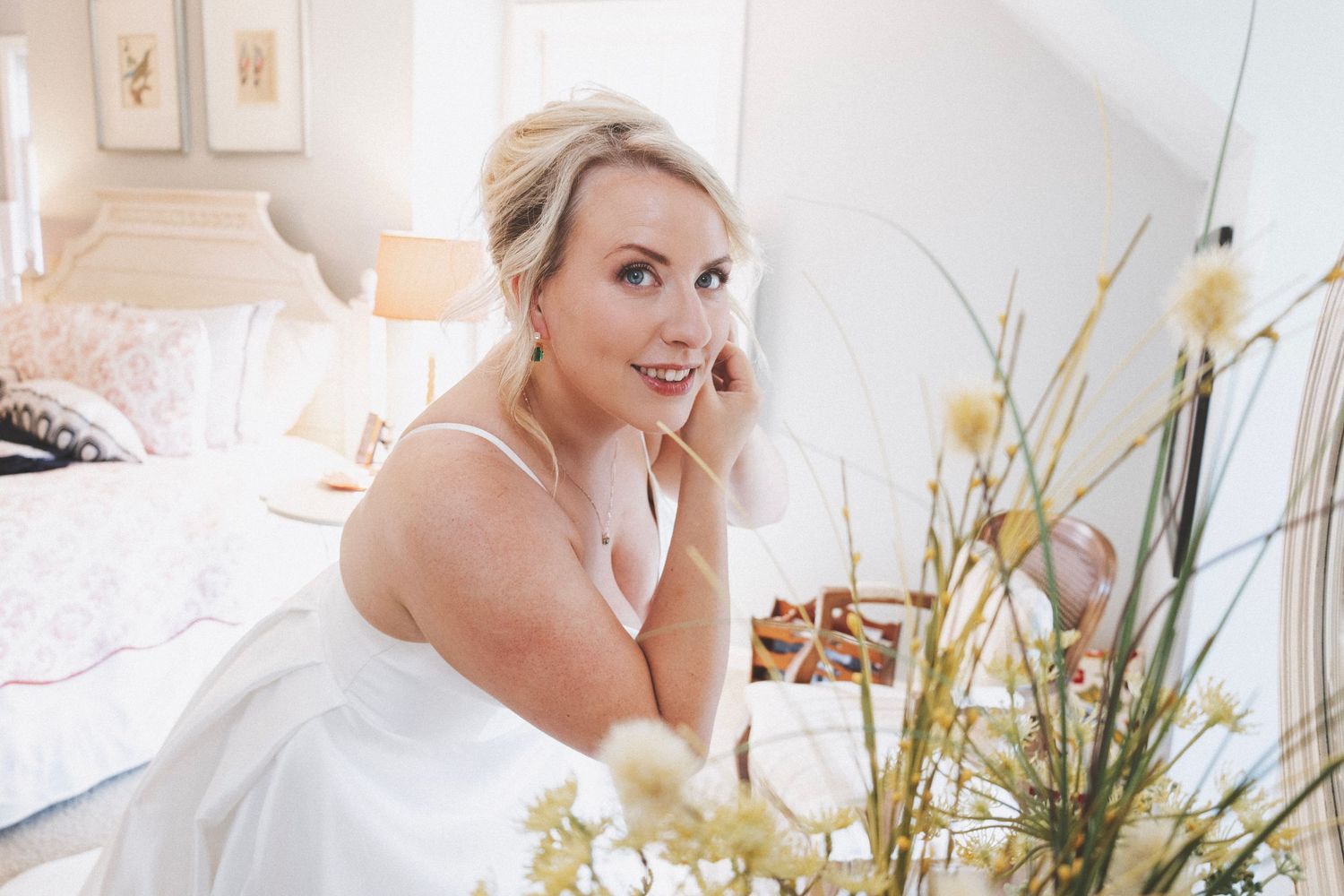 Elegant blonde bride in white gown sitting in a bright bedroom, smiling over her shoulder with fresh flowers in foreground.