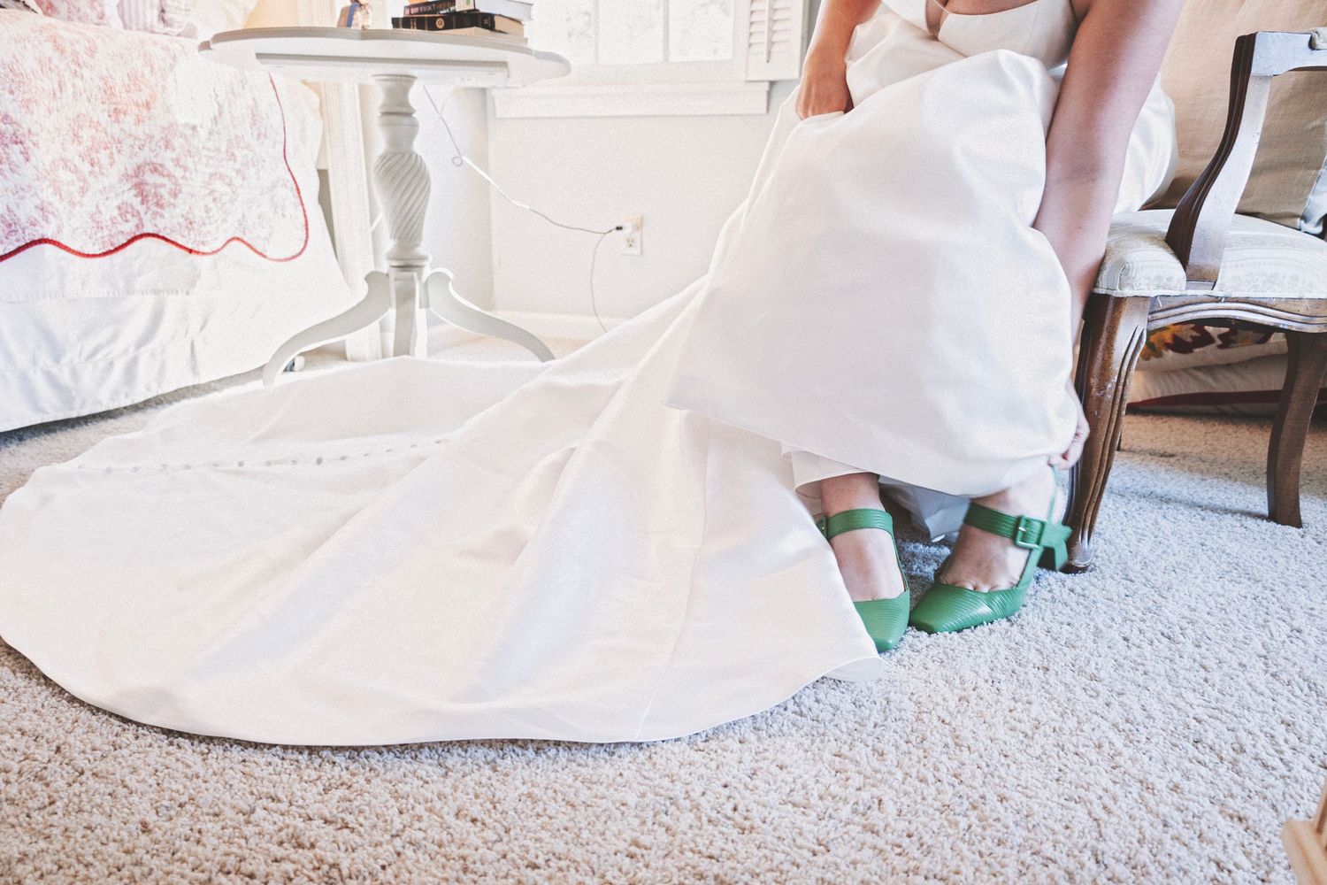 Bride in white wedding gown with green heels peeking out, standing in a decorated bedroom.