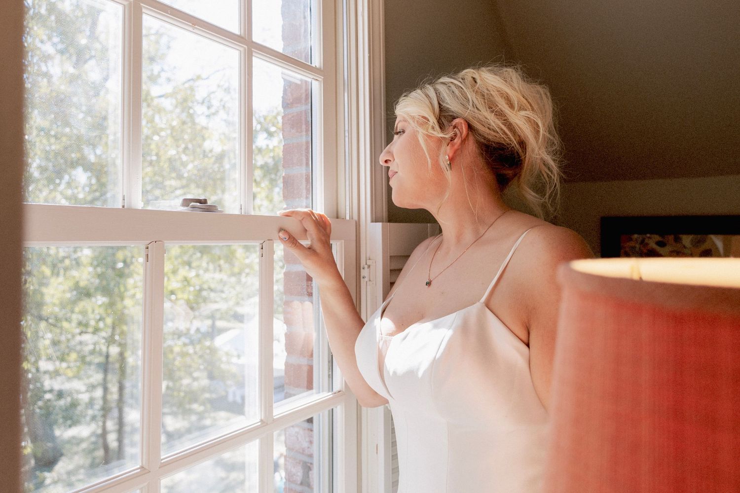 Blonde woman in white camisole gazing thoughtfully out a sunlit window beside a pink lamp.