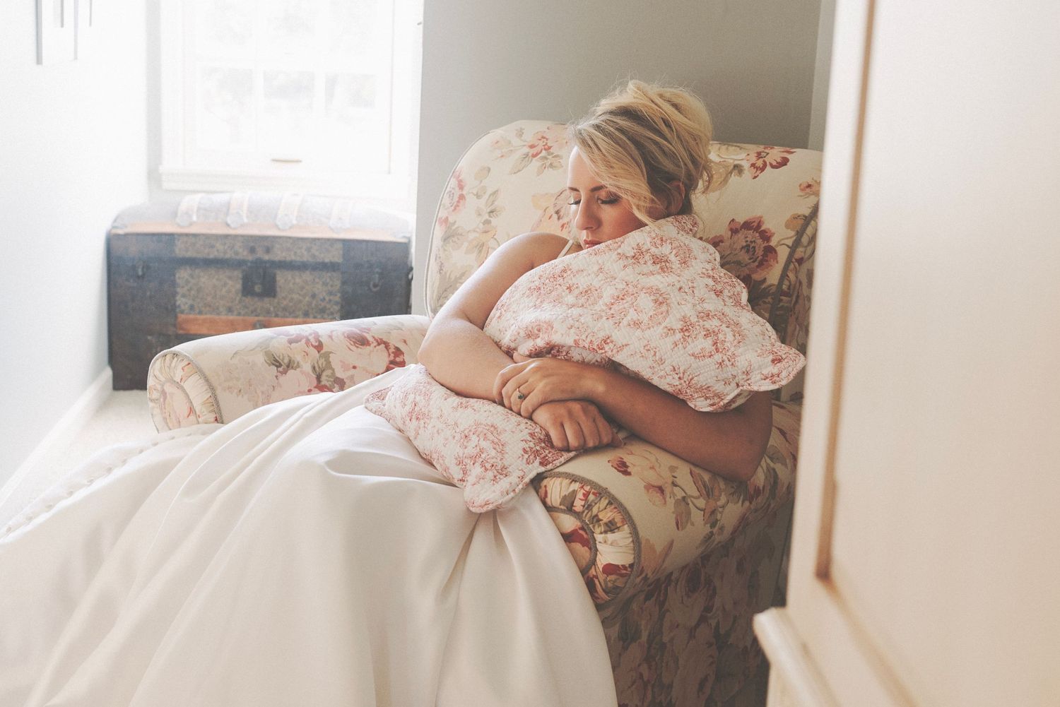 Blonde woman in a white dress hugging a floral pillow on an armchair in a bright, cozy room.