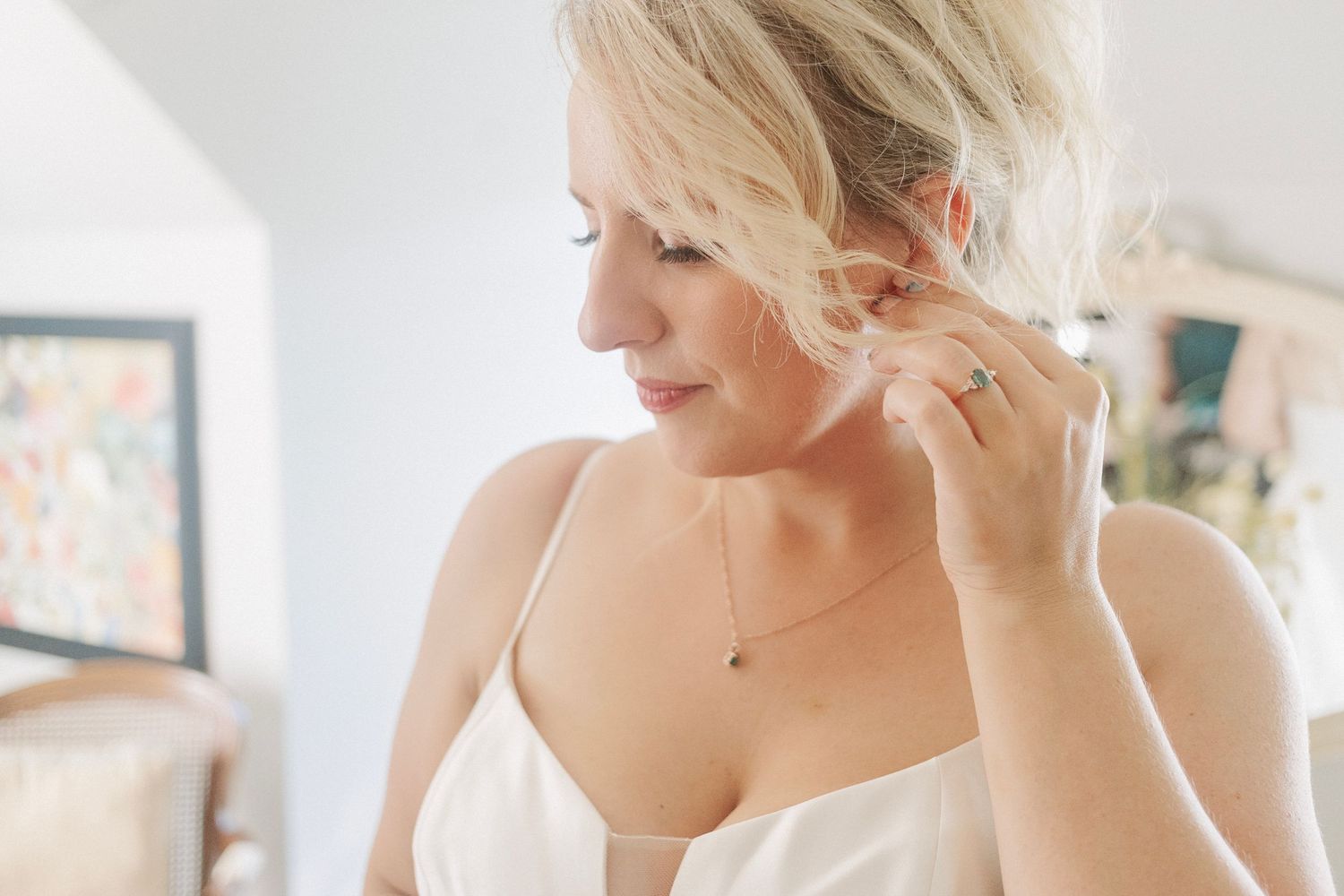 Blonde bride in white dress adjusting her earring, wearing a delicate necklace, in a softly lit room.