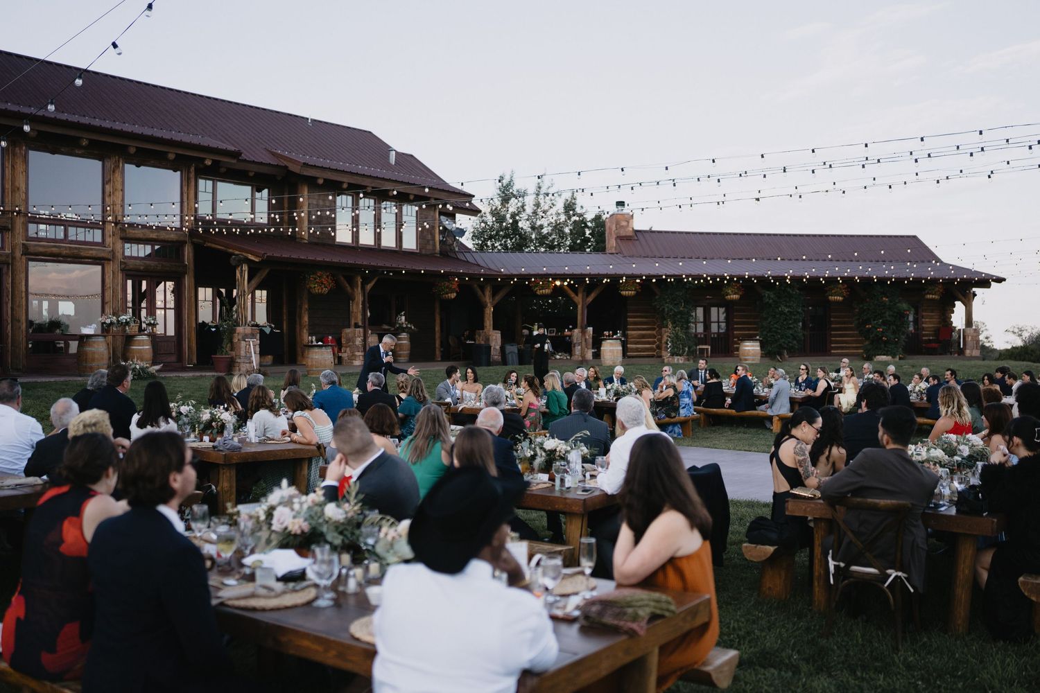 Outdoor wedding reception at rustic barn venue with string lights, wooden tables, and guests dining at dusk.