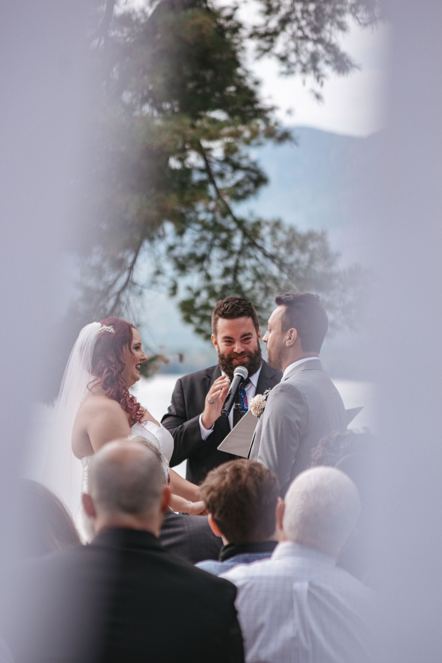 Outdoor wedding ceremony with bearded officiant reading vows to bride and groom before seated guests.