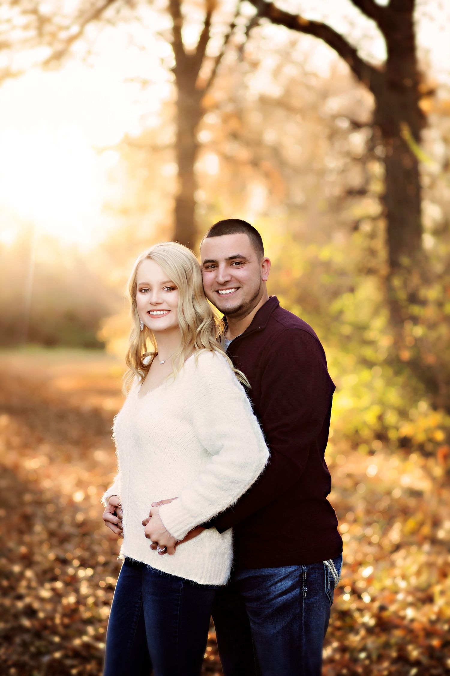 Couple poses outdoors in autumn park, woman in white sweater cradling baby bump, man in burgundy top embracing her from behind.