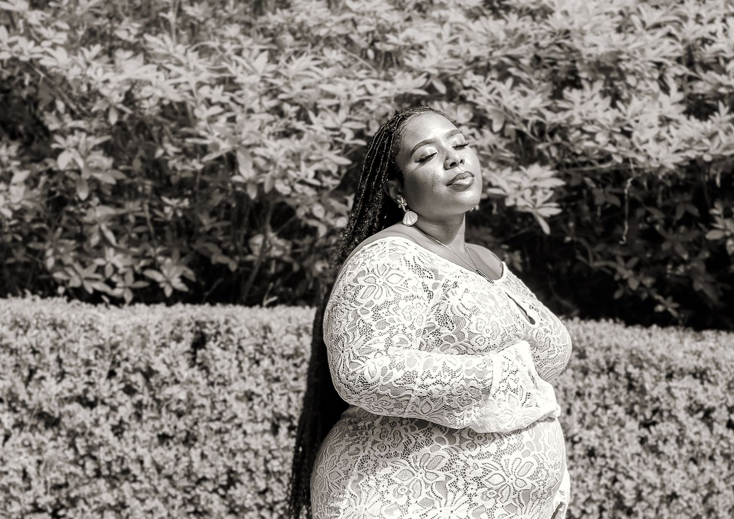 Black and white portrait of a pregnant woman in lace dress posing confidently outdoors near lush green hedges.