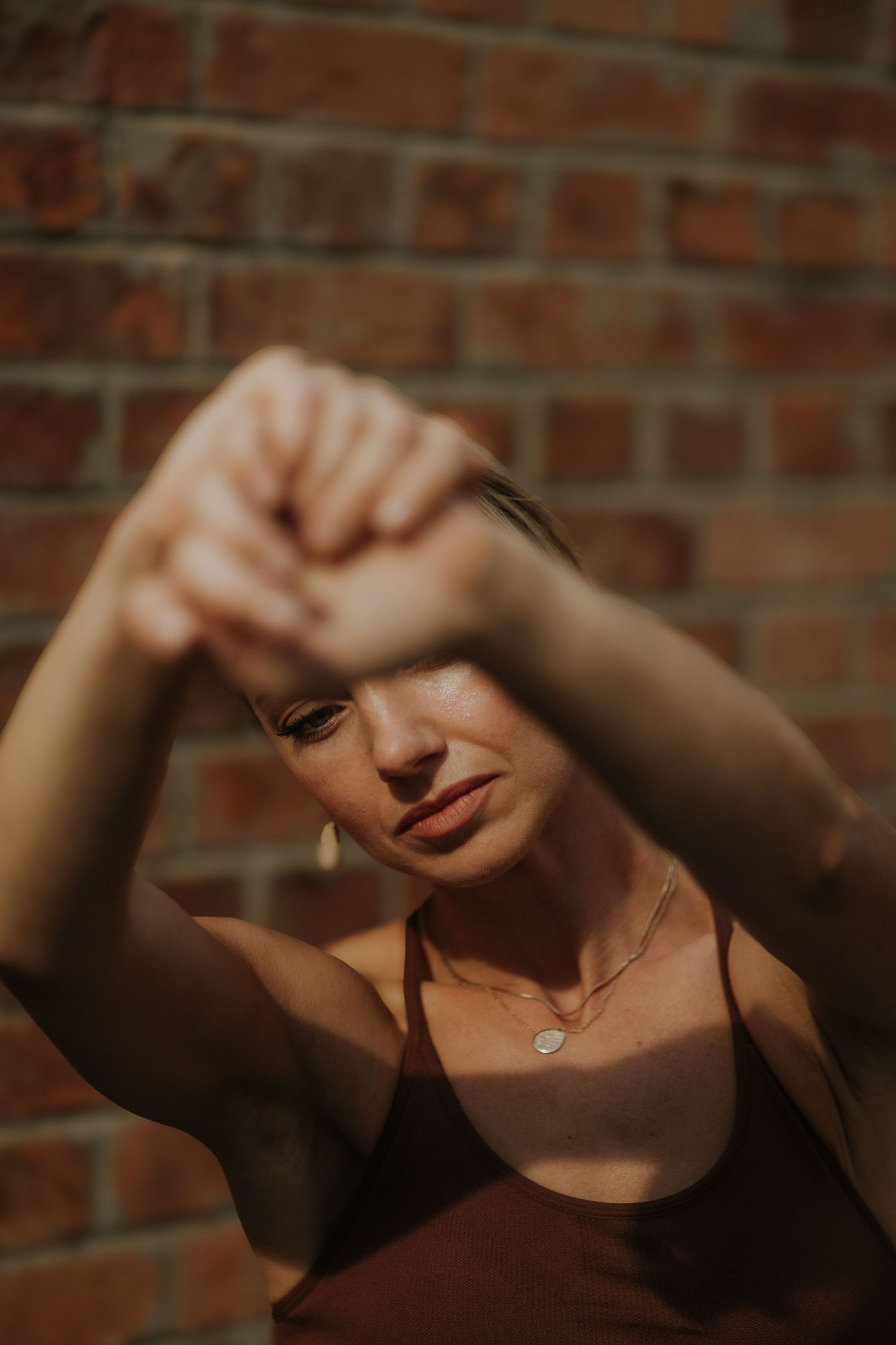 Woman in brown top poses with arms raised near brick wall, sunlight casting dramatic shadows on her face.