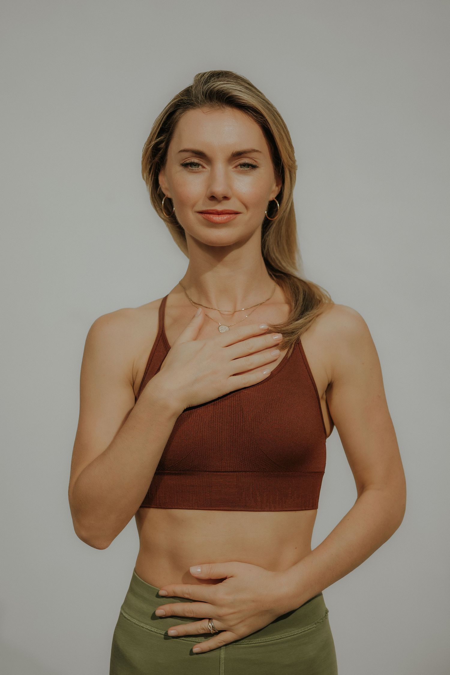 Woman in brown sports bra and olive leggings poses with hands on chest and stomach against gray background.