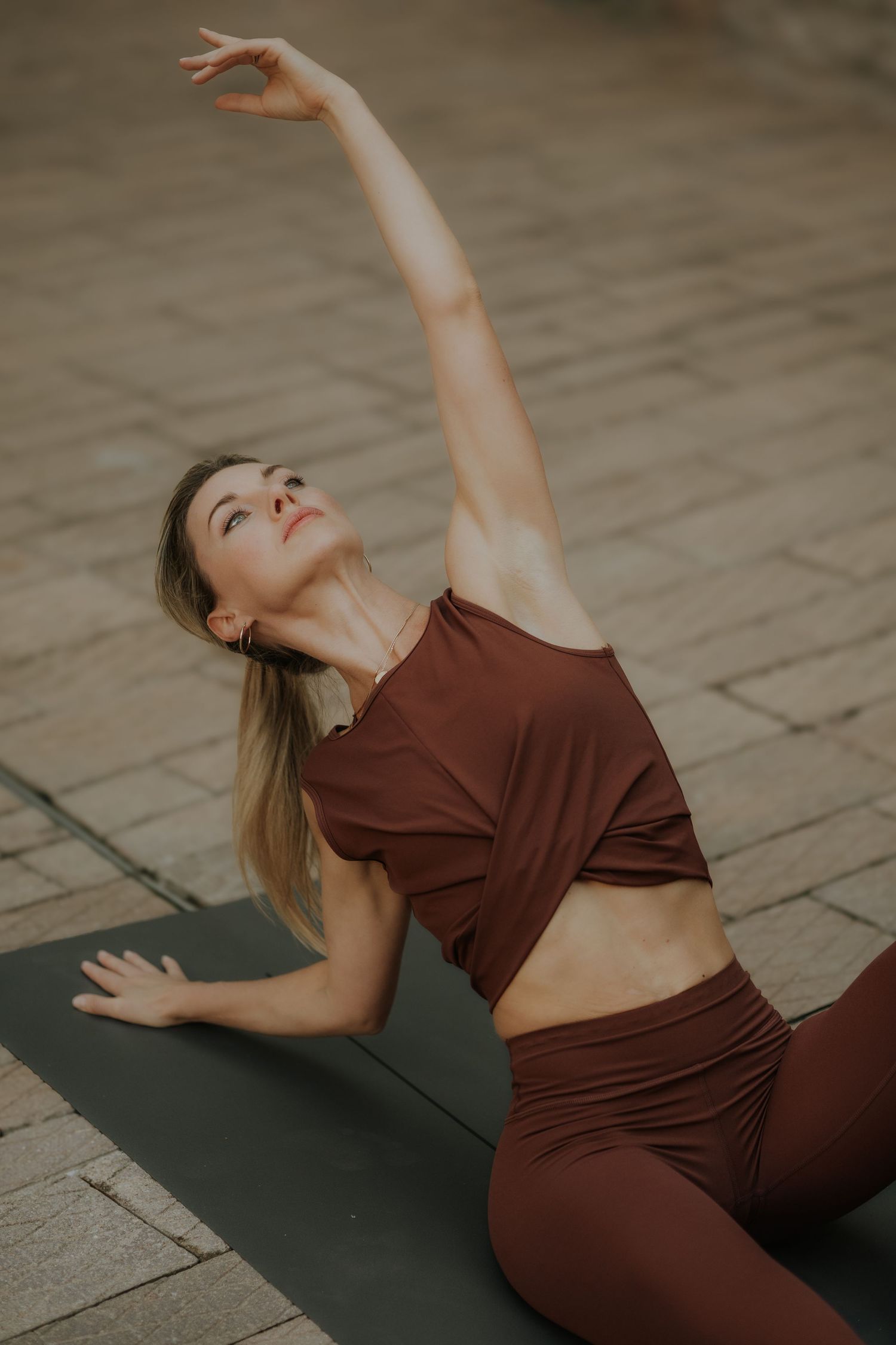 Woman in brown activewear doing a side stretch yoga pose on a mat outdoors on a cobblestone surface.