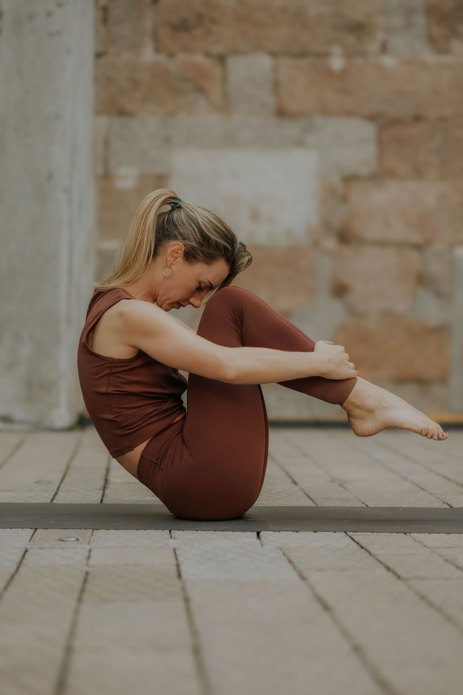 Woman in brown activewear performing a seated yoga pose on a mat outdoors against a stone wall.
