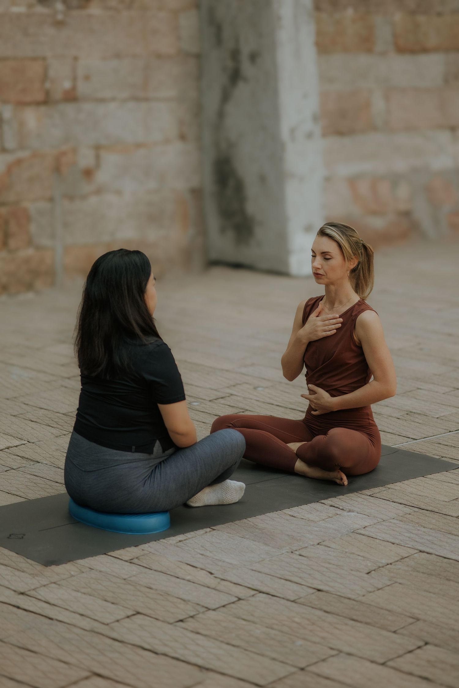 Two women sit cross-legged on yoga mats outdoors, practicing mindfulness breathing exercises together near a stone wall.