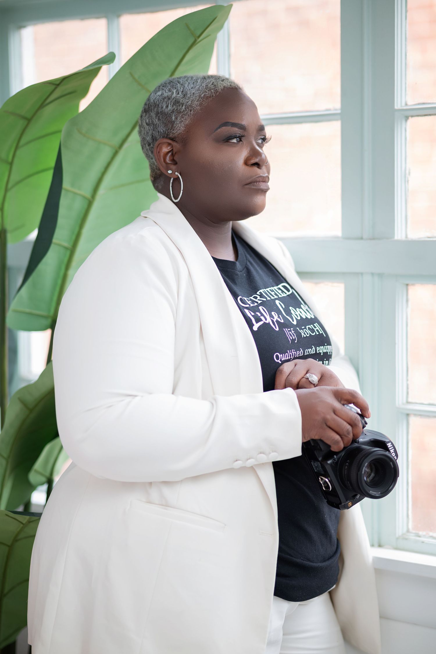 A Black woman photographer in a white blazer holds a camera, standing near a window with tropical plant decor.
