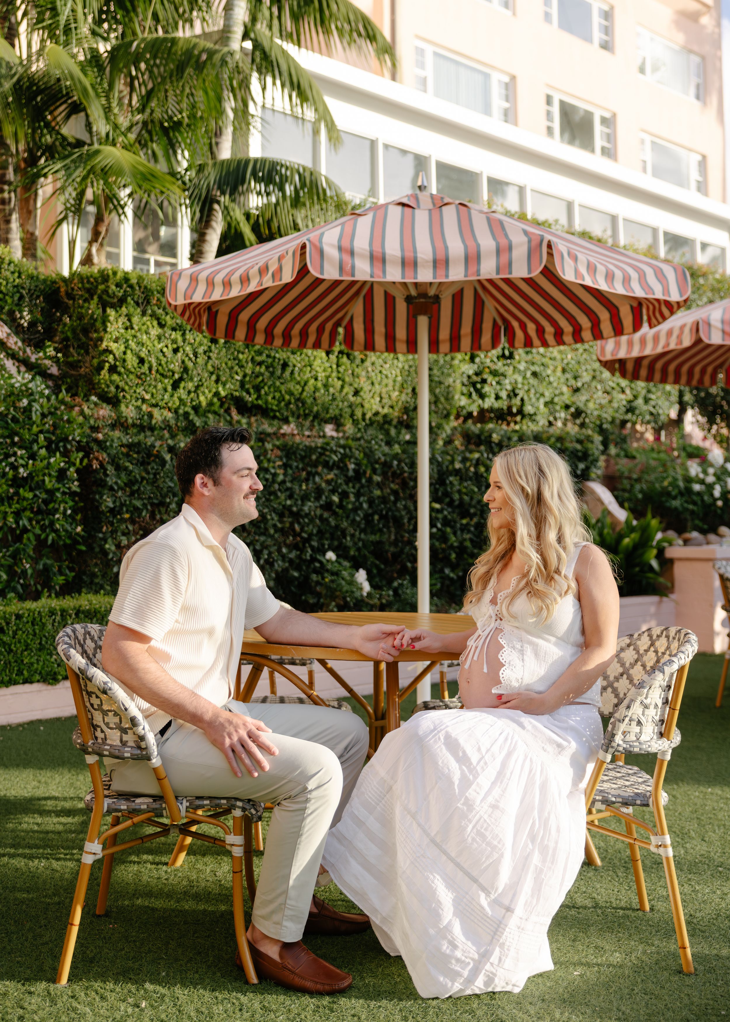 Couple sitting at outdoor cafe table under striped umbrella on green lawn with tropical hotel backdrop.