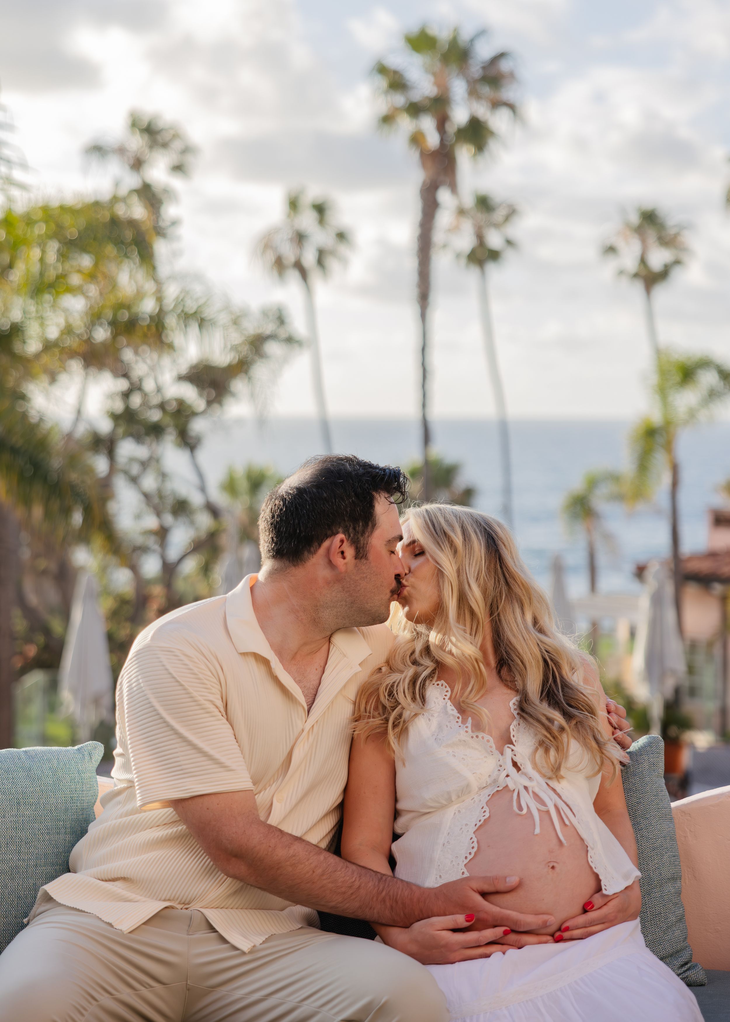 Couple sharing a kiss during maternity photo shoot with palm trees and ocean in the background.