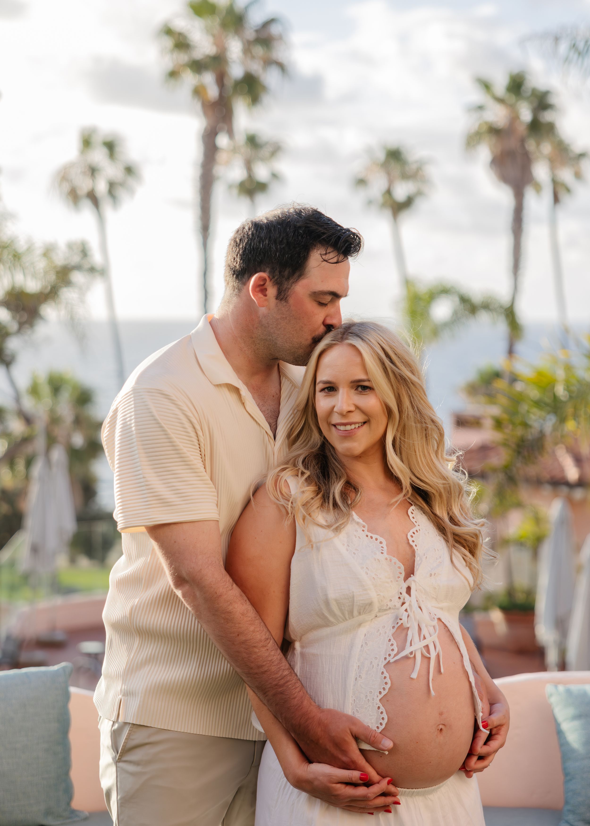 A couple poses for a maternity photo outdoors with palm trees, the man kissing her head while cradling her baby bump.