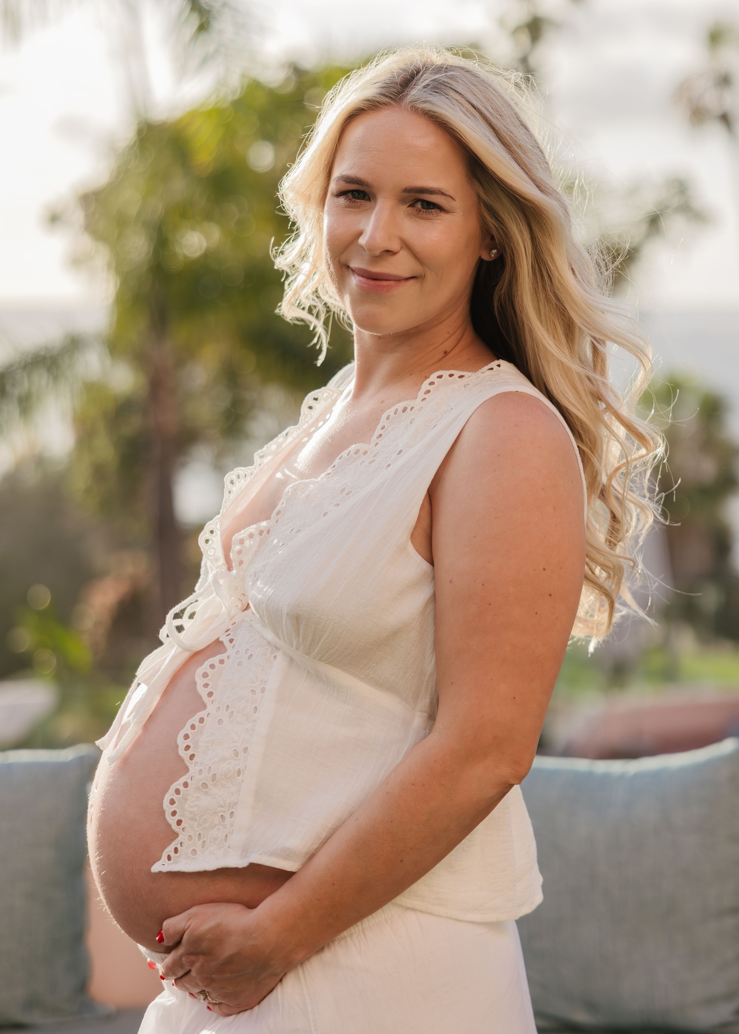 Smiling pregnant woman with long blonde hair wearing a white lace top, cradling her baby bump outdoors.