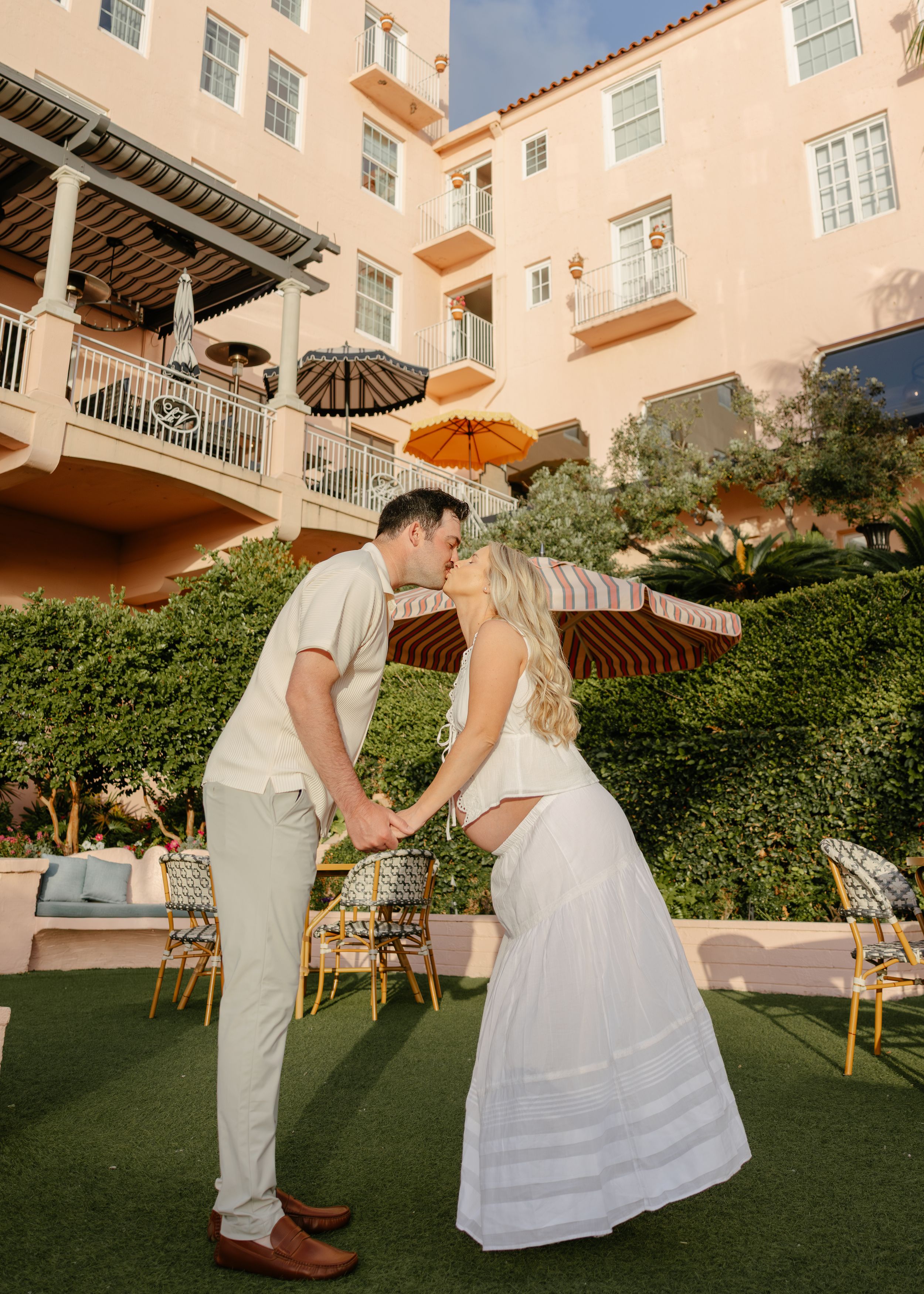 Couple kissing and holding hands in front of a pink Spanish-style hotel courtyard with striped umbrellas.