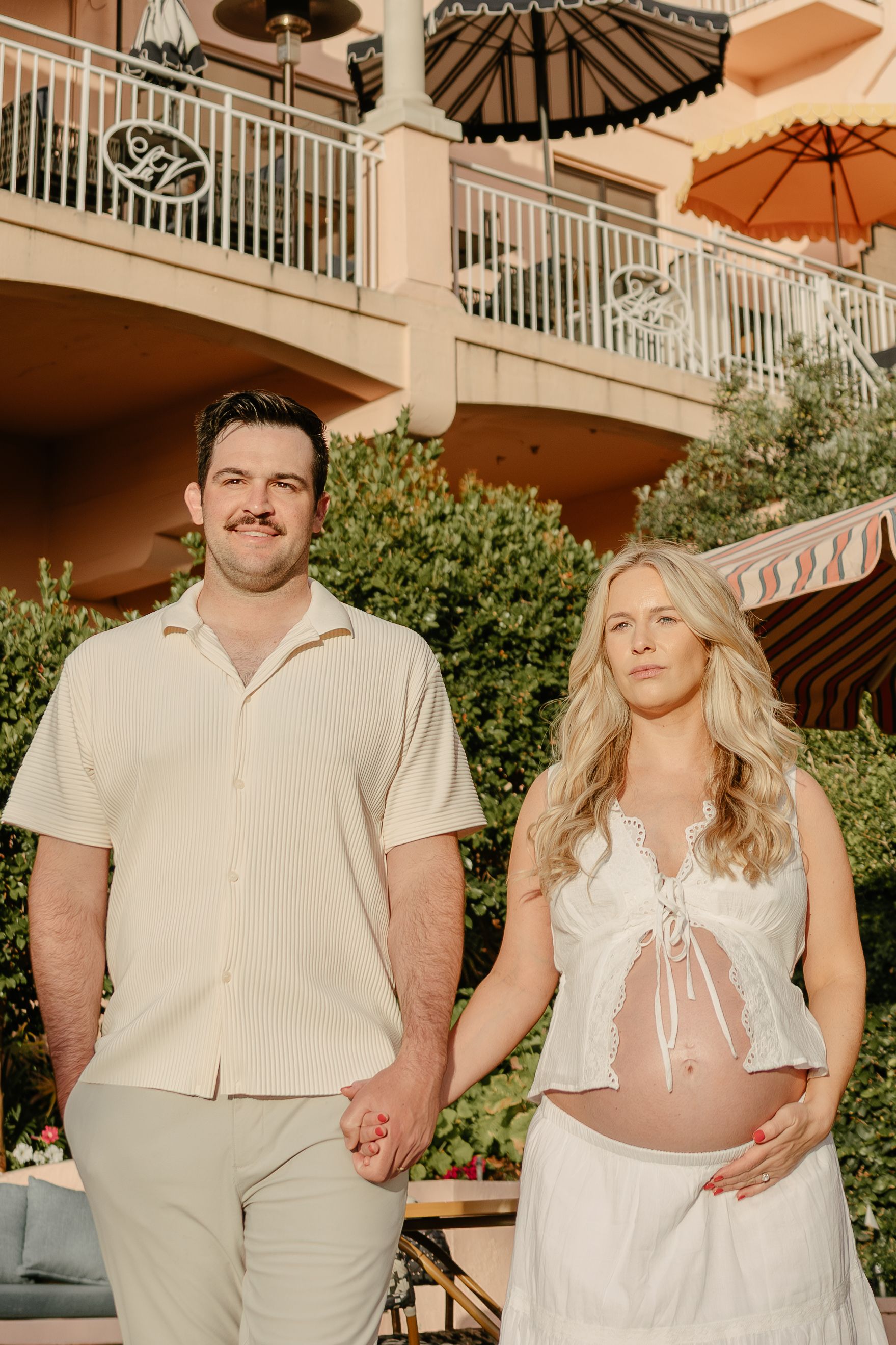 Pregnant couple holding hands outdoors at a resort, woman in white maternity outfit, man in cream shirt and pants.
