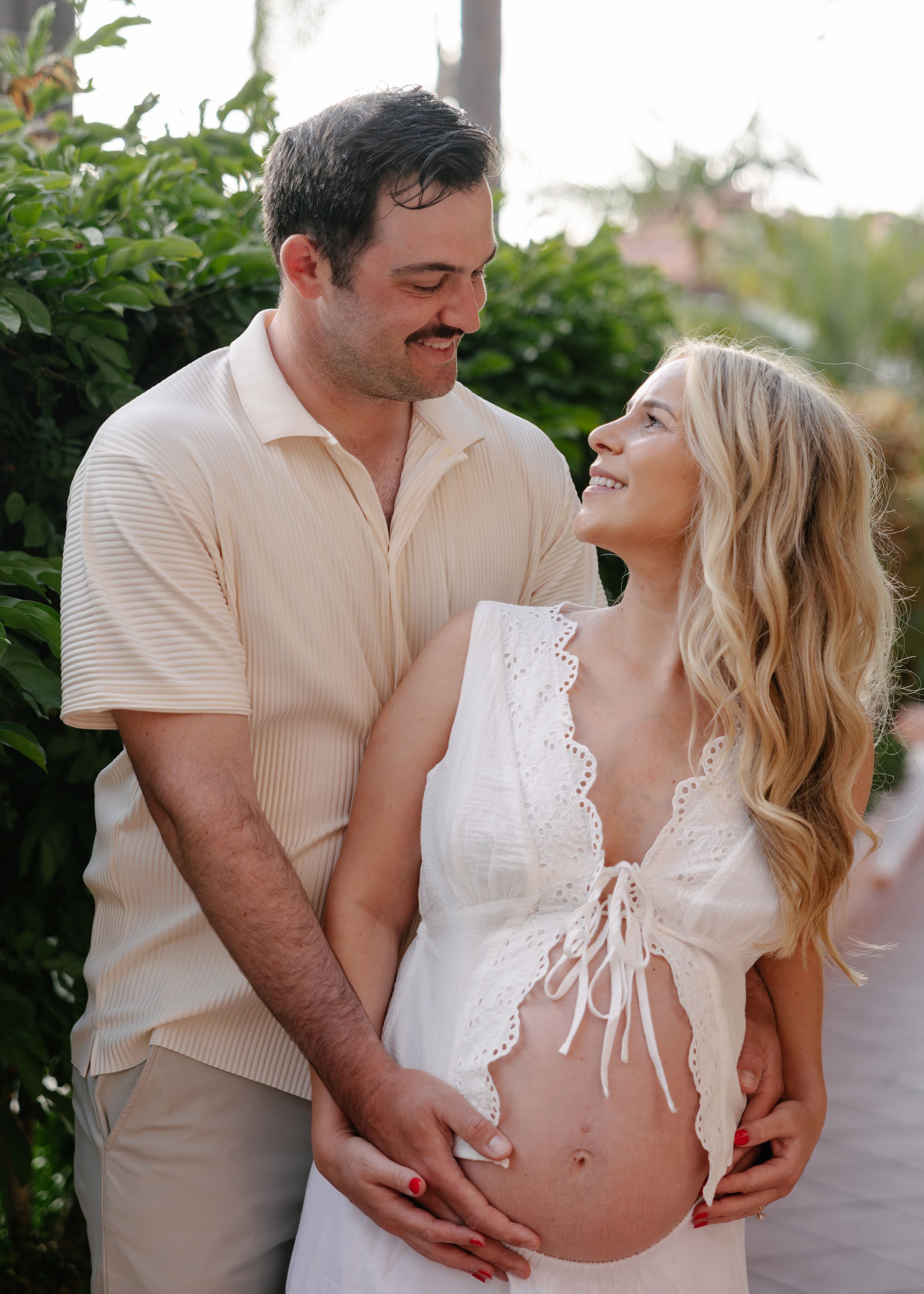 Happy couple sharing a tender moment outdoors, woman in white maternity outfit showing baby bump, man in beige polo shirt.