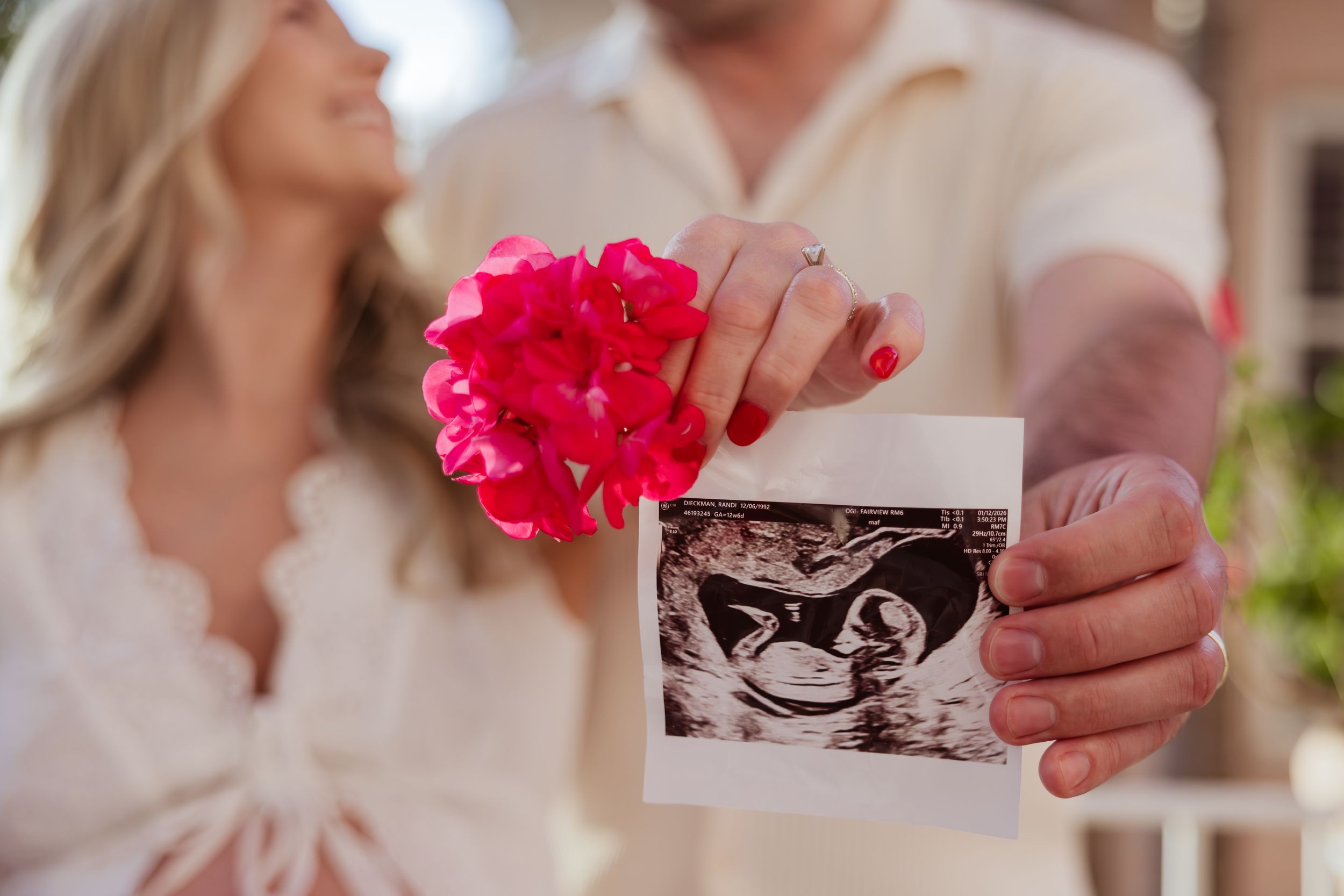 Couple holding ultrasound photo and pink flowers, announcing pregnancy with smiles in a romantic outdoor setting.
