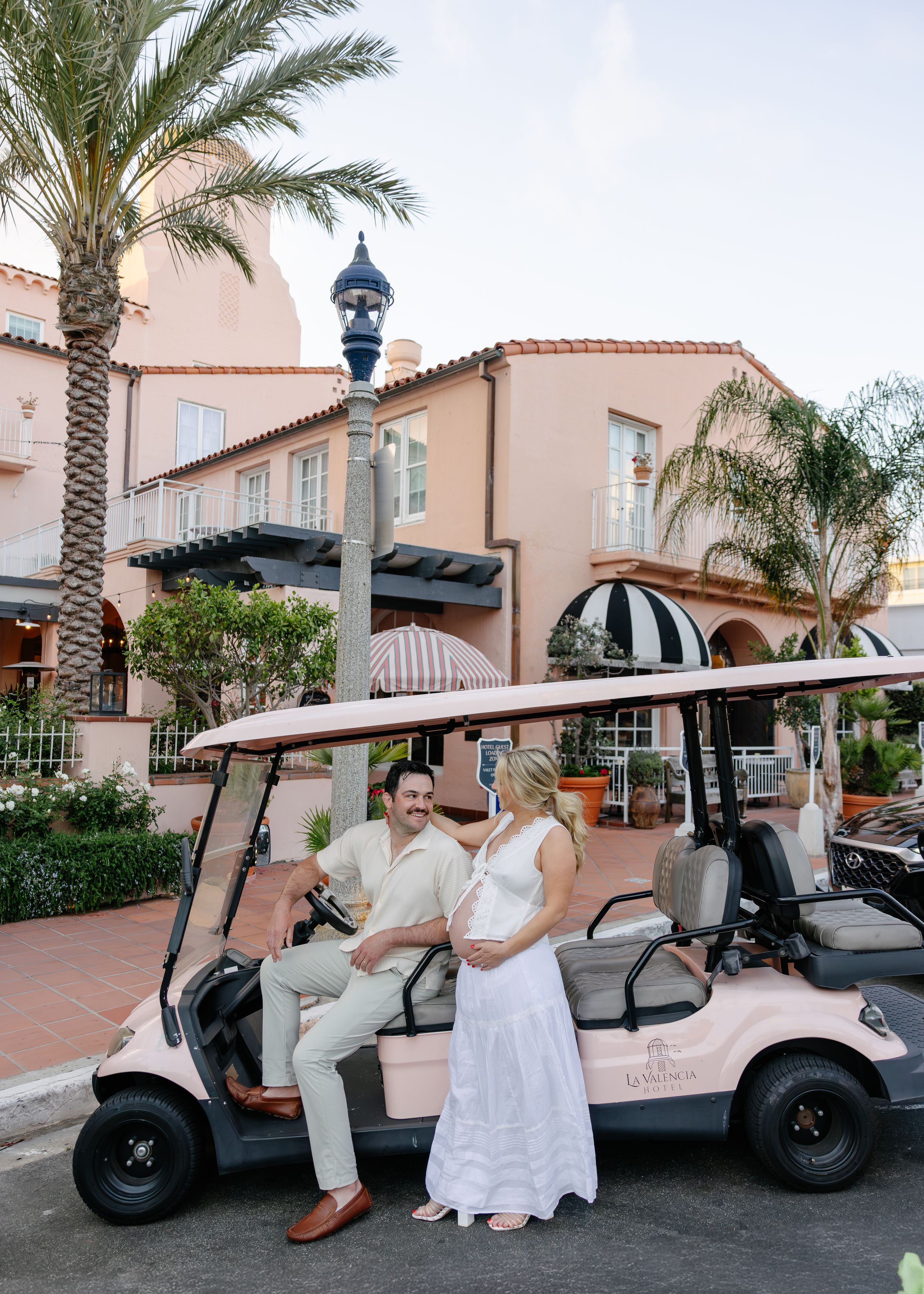 Couple in wedding attire posing with a pink golf cart outside a Spanish-style resort with palm trees.