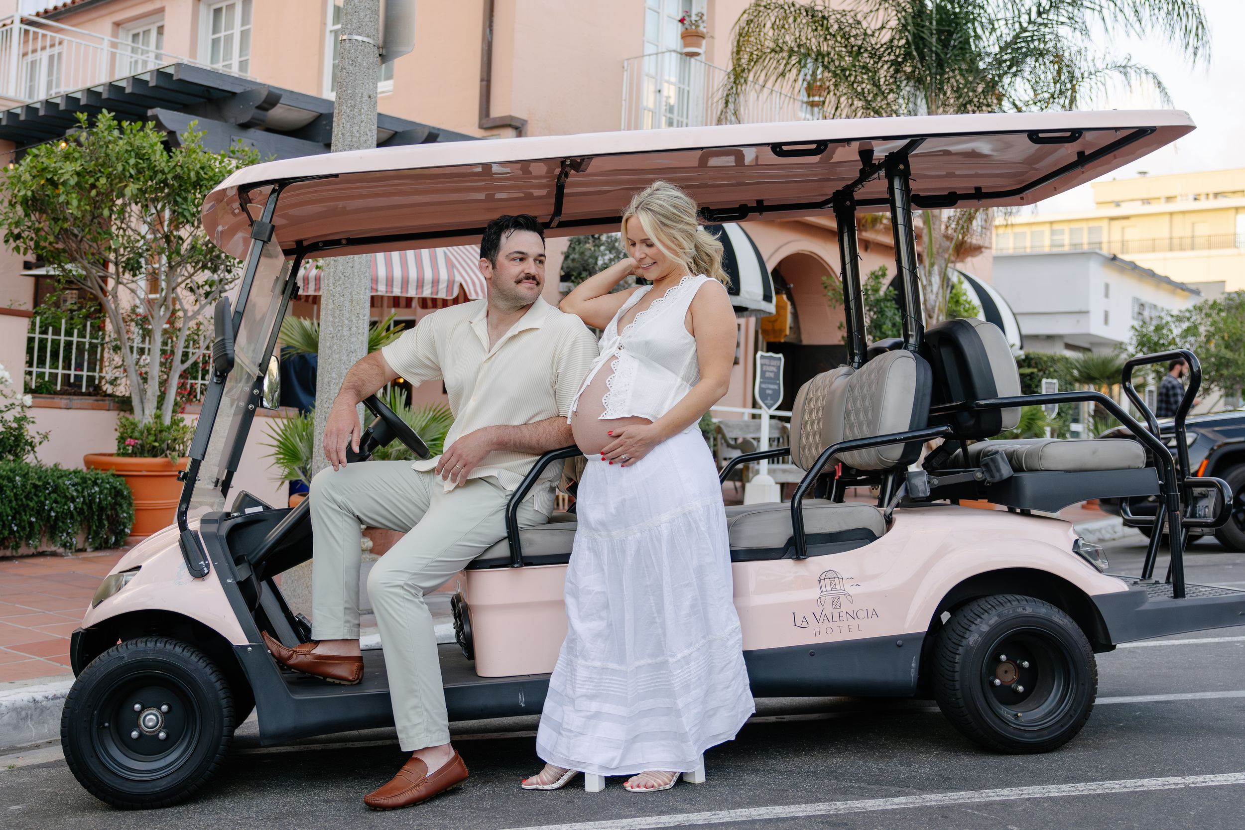 Pregnant couple poses beside a pink golf cart on a sunny street, woman in white dress, man in beige outfit.