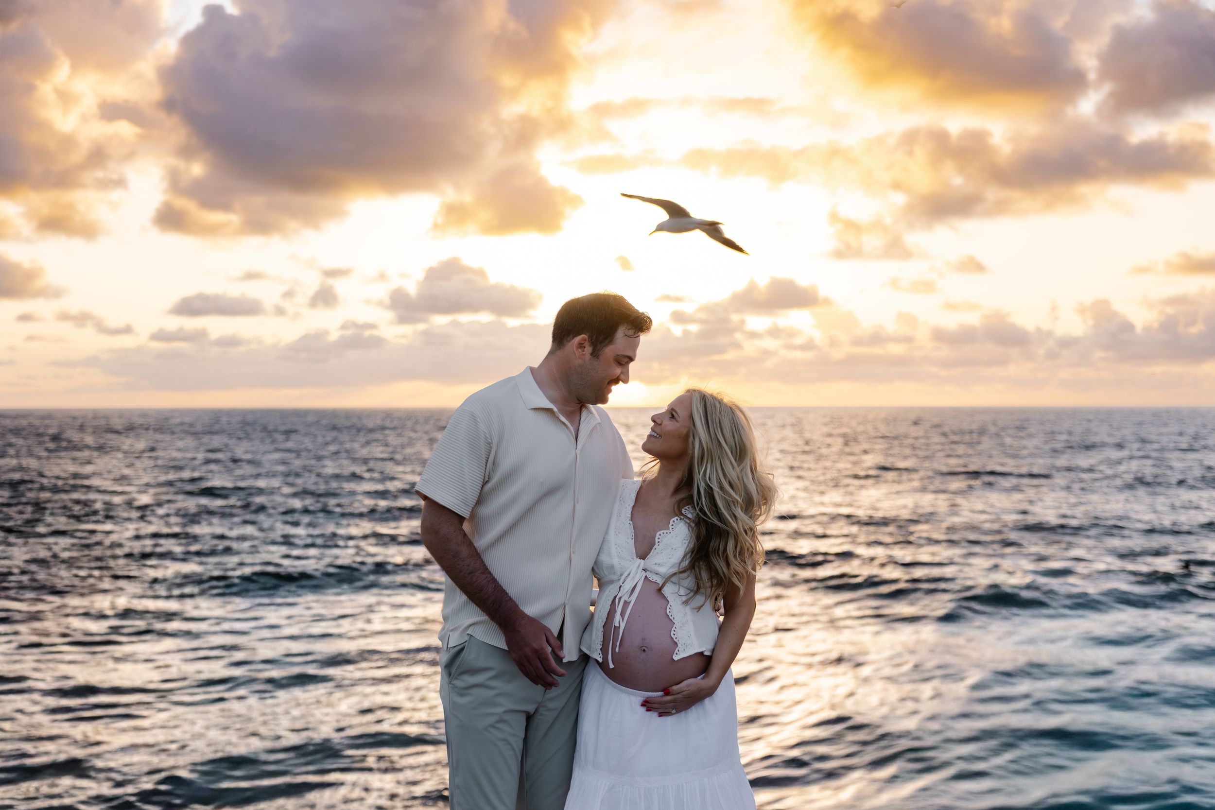 Couple shares a romantic moment at sunset by the ocean, with a seagull soaring above during a maternity photo shoot.