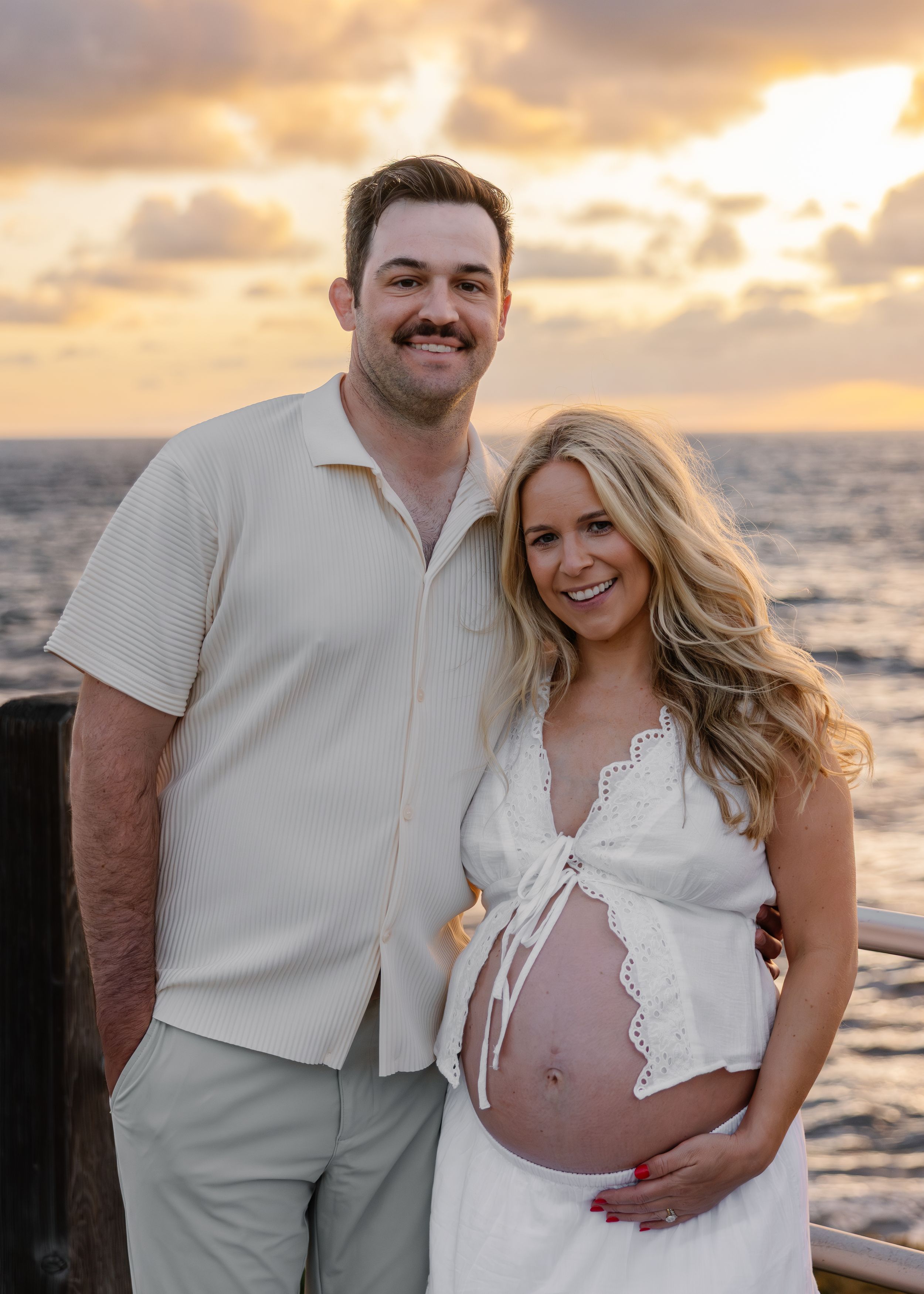 Couple posing at sunset by the ocean, woman in white dress cradling baby bump, man in beige shirt and pants smiling.
