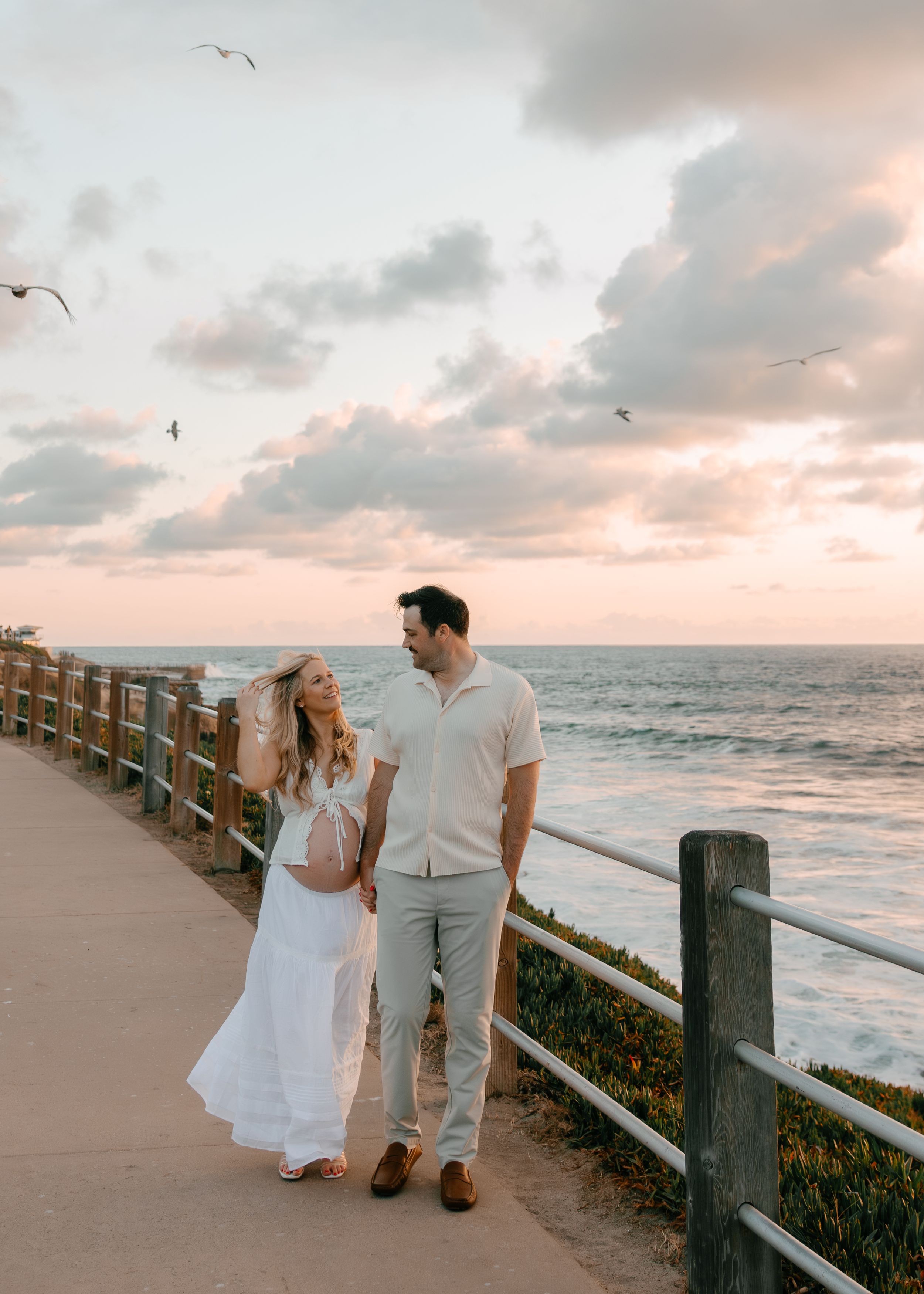 Couple walking along ocean boardwalk at sunset, woman in white dress holding hat, man in cream shirt and gray pants.