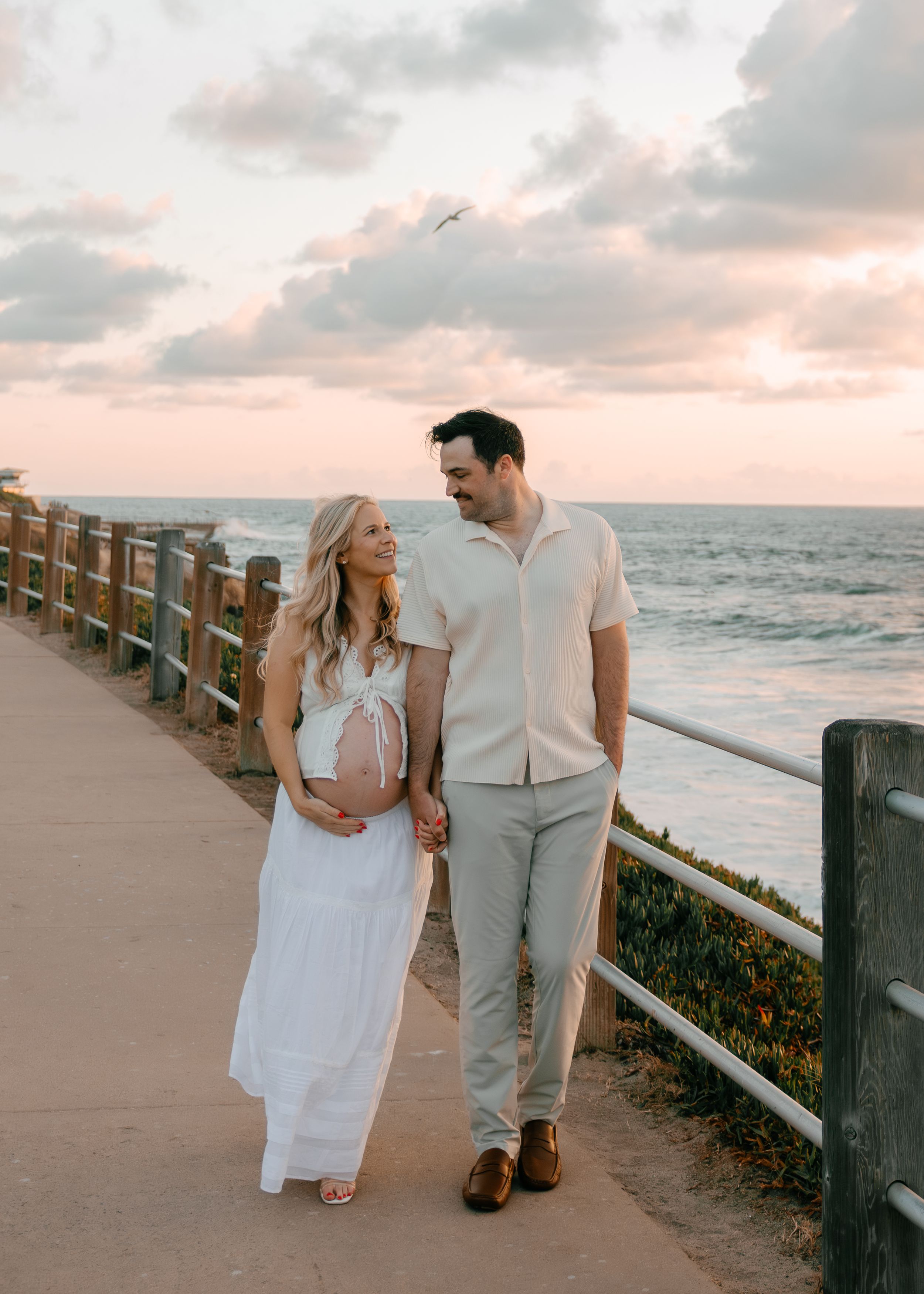 Pregnant couple holding hands on coastal walkway at sunset, woman in white maxi dress, man in light casual outfit.
