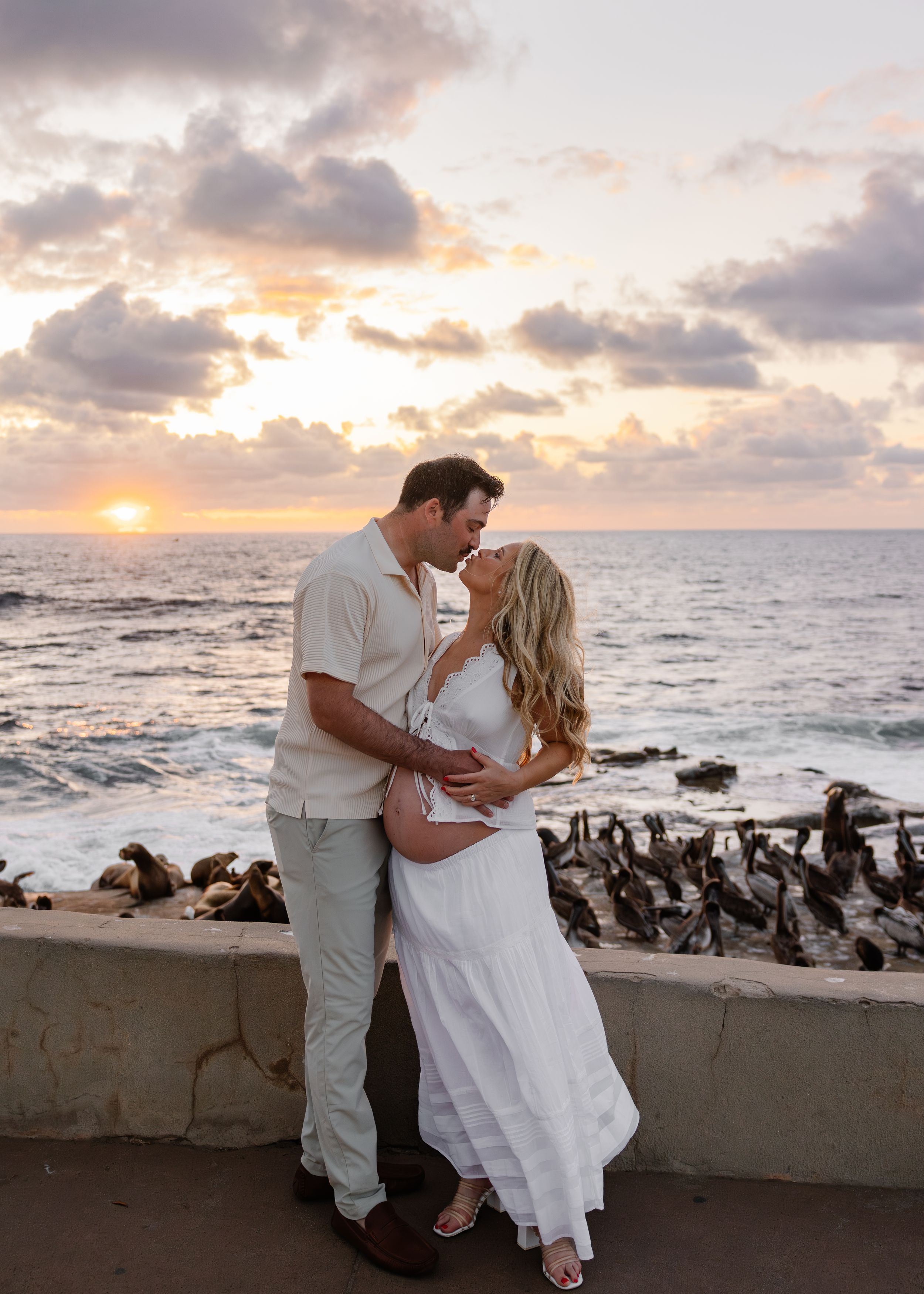 Couple sharing a romantic kiss at sunset by the ocean, woman in white dress with baby bump, man in beige outfit.