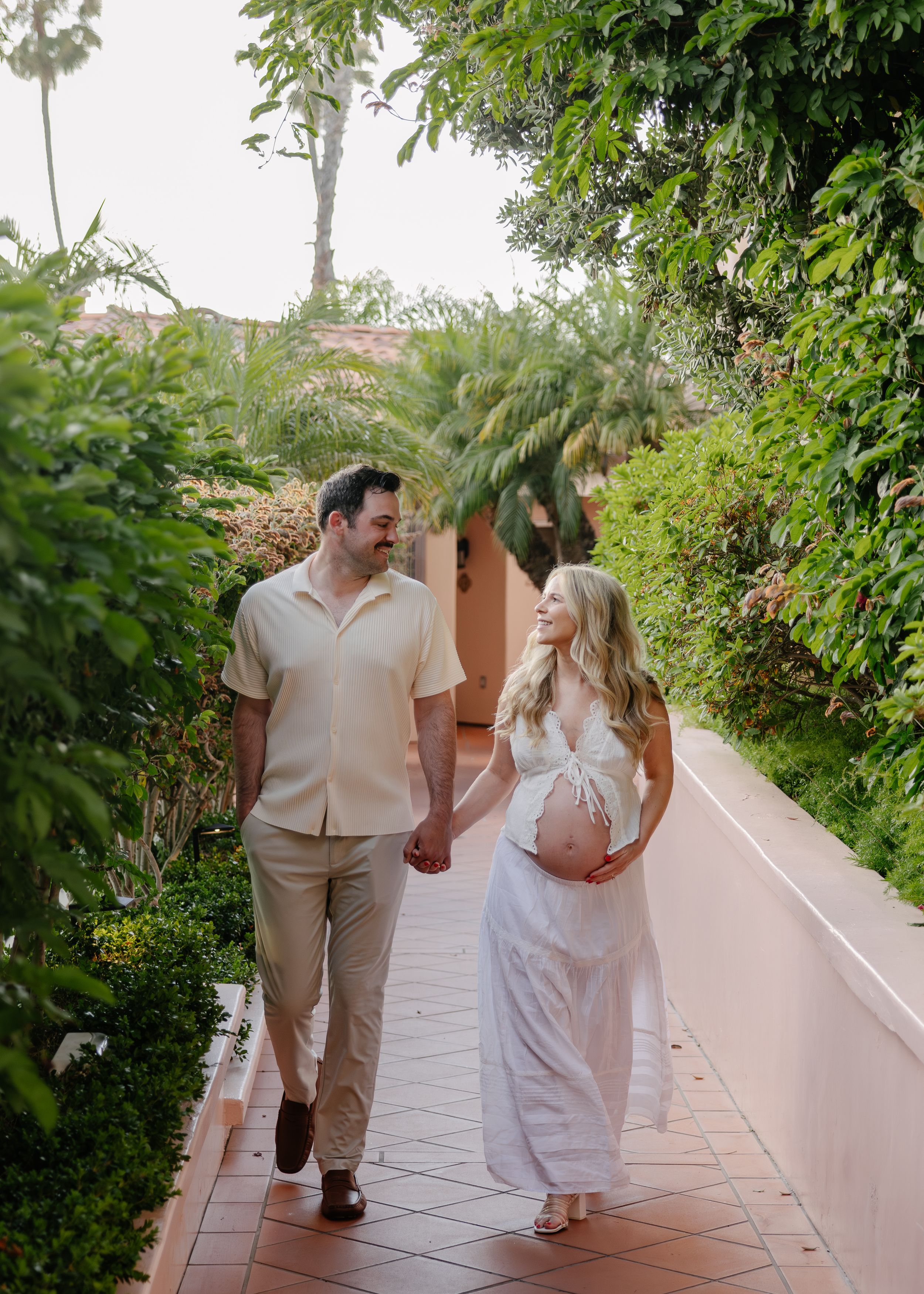 Pregnant couple holding hands walking along a tropical garden path, woman in white dress cradling baby bump.