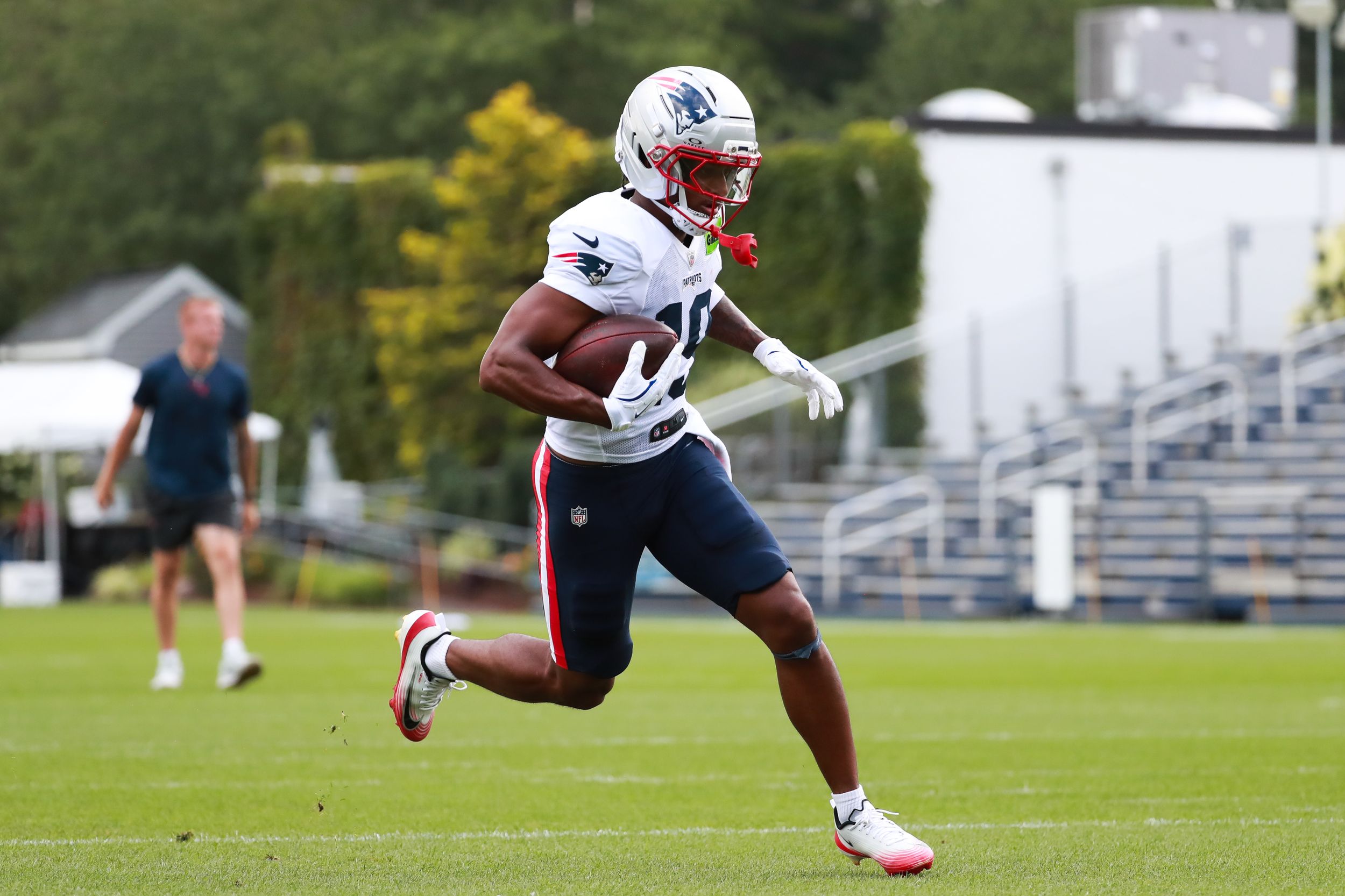 New England Patriots player runs with football during outdoor practice drill in white uniform.