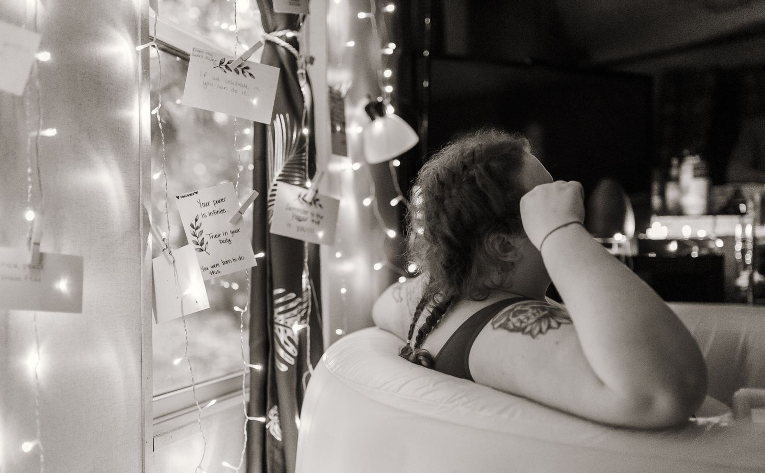 Black and white photo of a woman relaxing near fairy lights and pinned notes in a cozy decorated room.