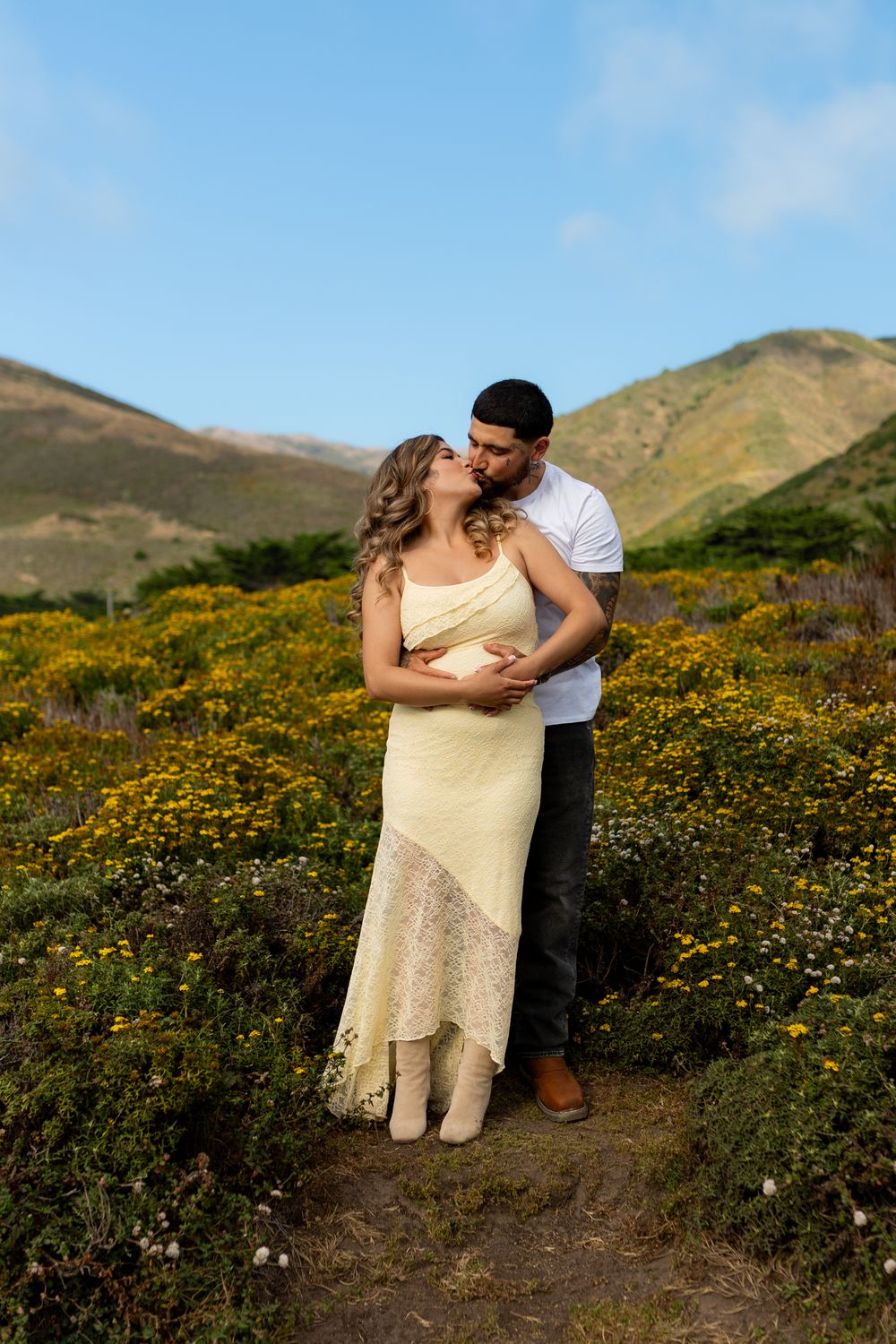 Couple kissing in a field of yellow wildflowers with green hills and blue sky in the background.