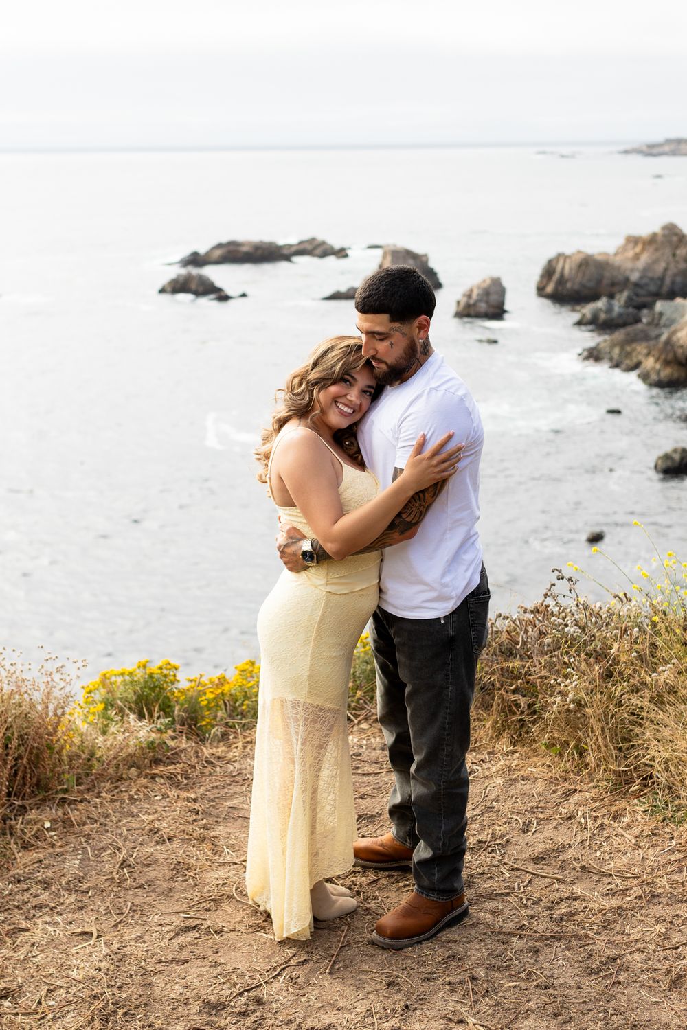 Couple embracing on coastal cliffside, woman in yellow dress, man in white shirt, rocky ocean backdrop.