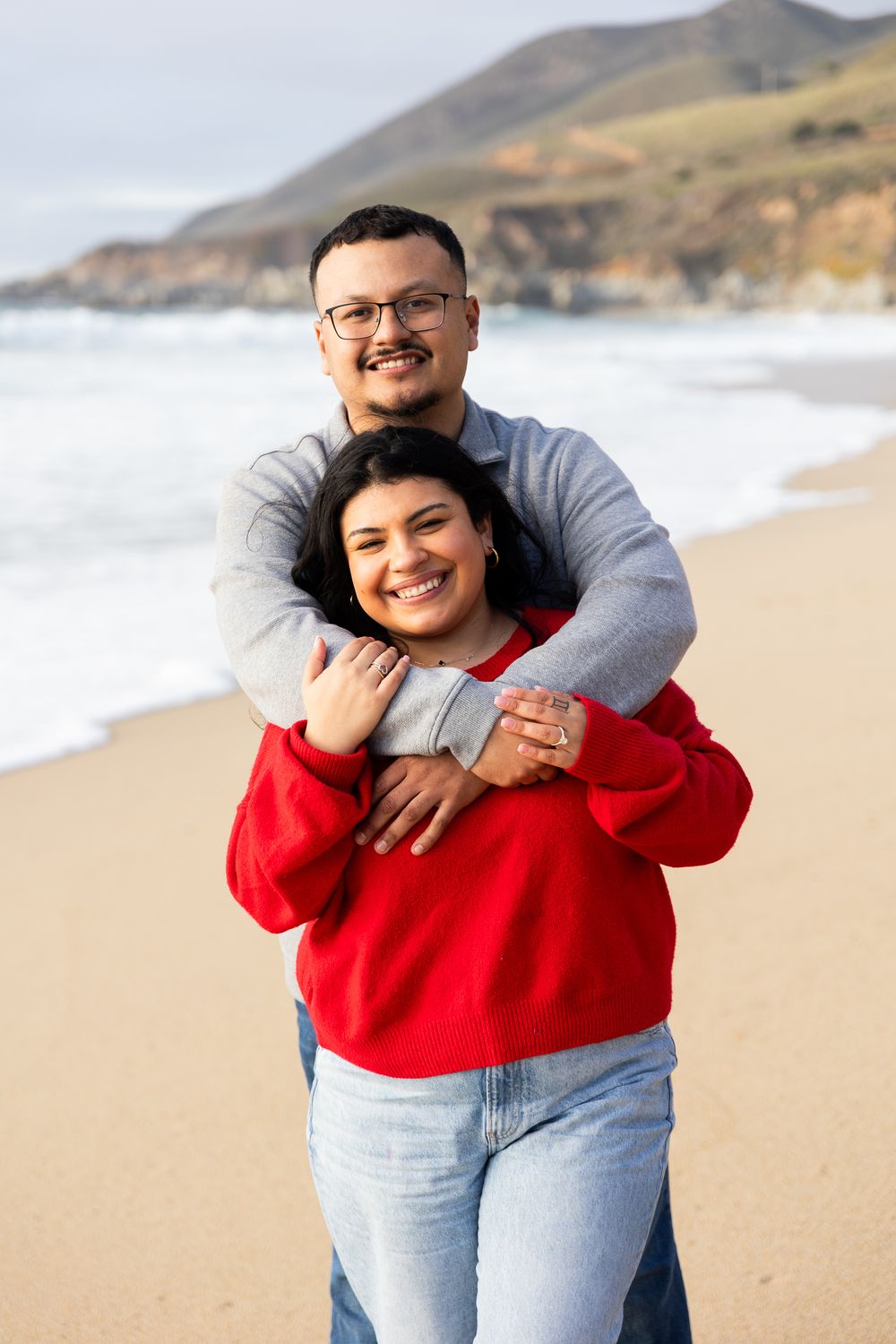 Happy couple embracing on a sandy beach with ocean waves and hills in the background.