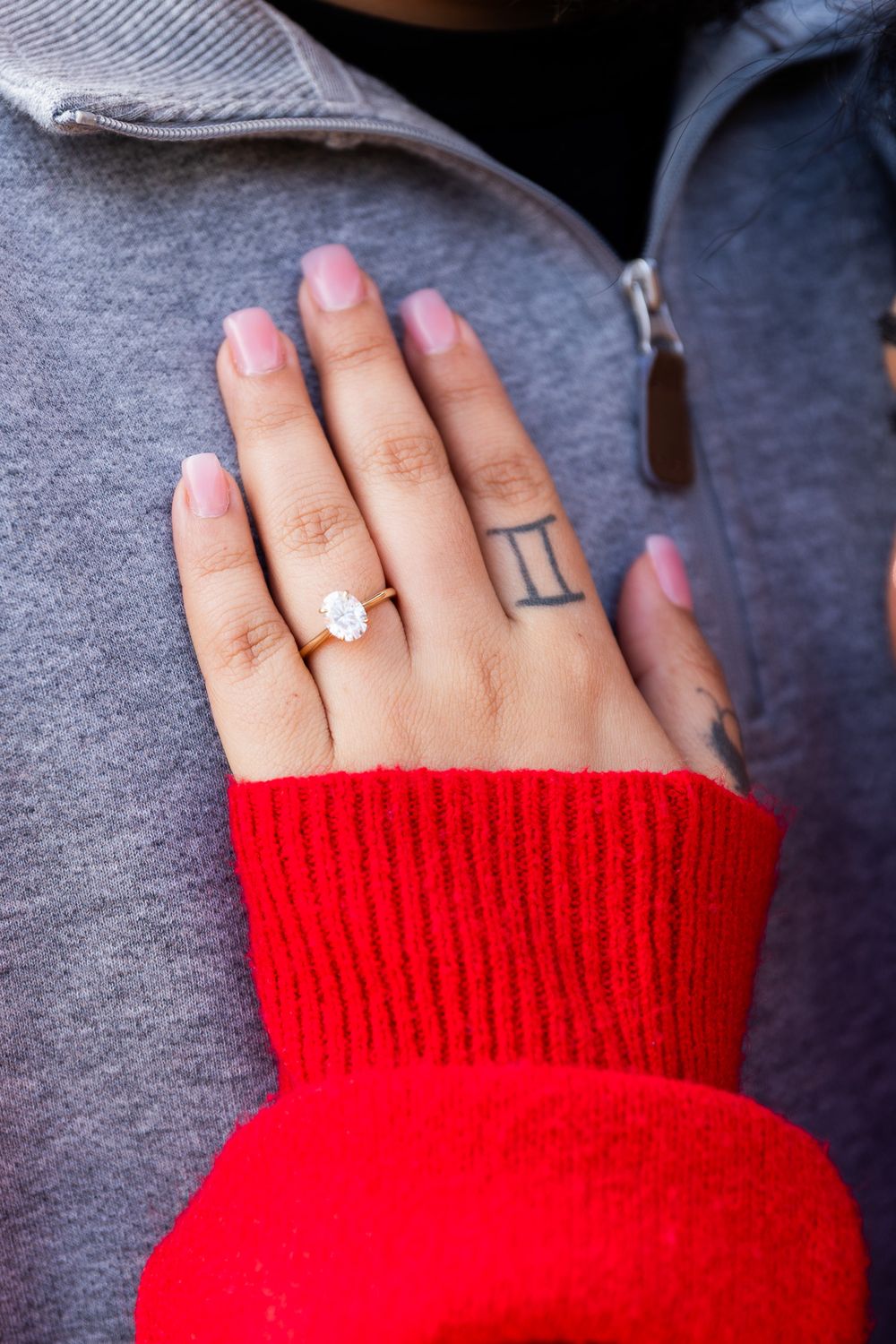 Close-up of hand wearing diamond engagement ring, red sweater sleeve, Gemini tattoo on finger.