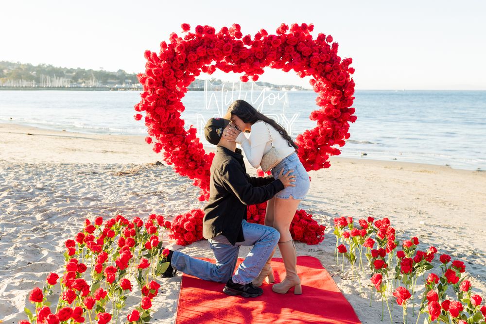Couple kissing during romantic beach proposal surrounded by red roses and heart-shaped floral arch at sunset.