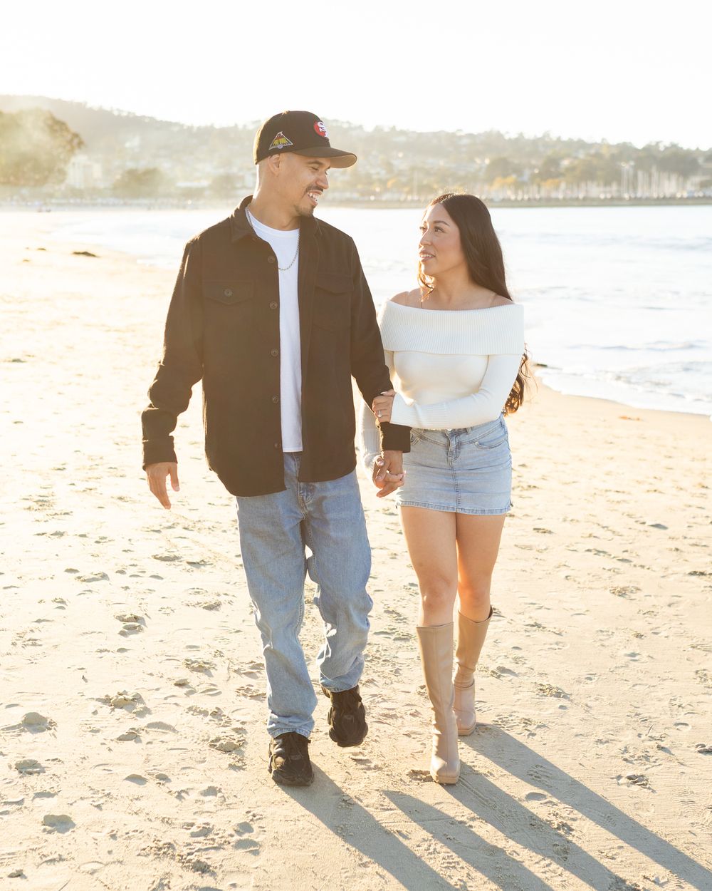 Couple holding hands walking on a sunny beach, man in black jacket and jeans, woman in white top and denim shorts.