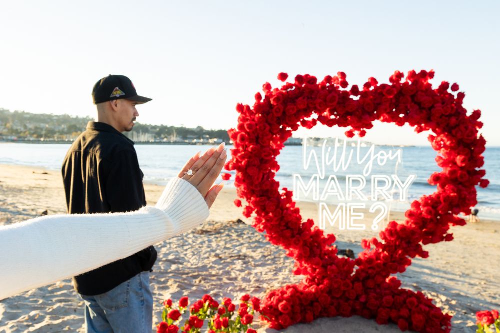 Man proposing to partner on beach with large red rose heart and Will You Marry Me sign at sunset.